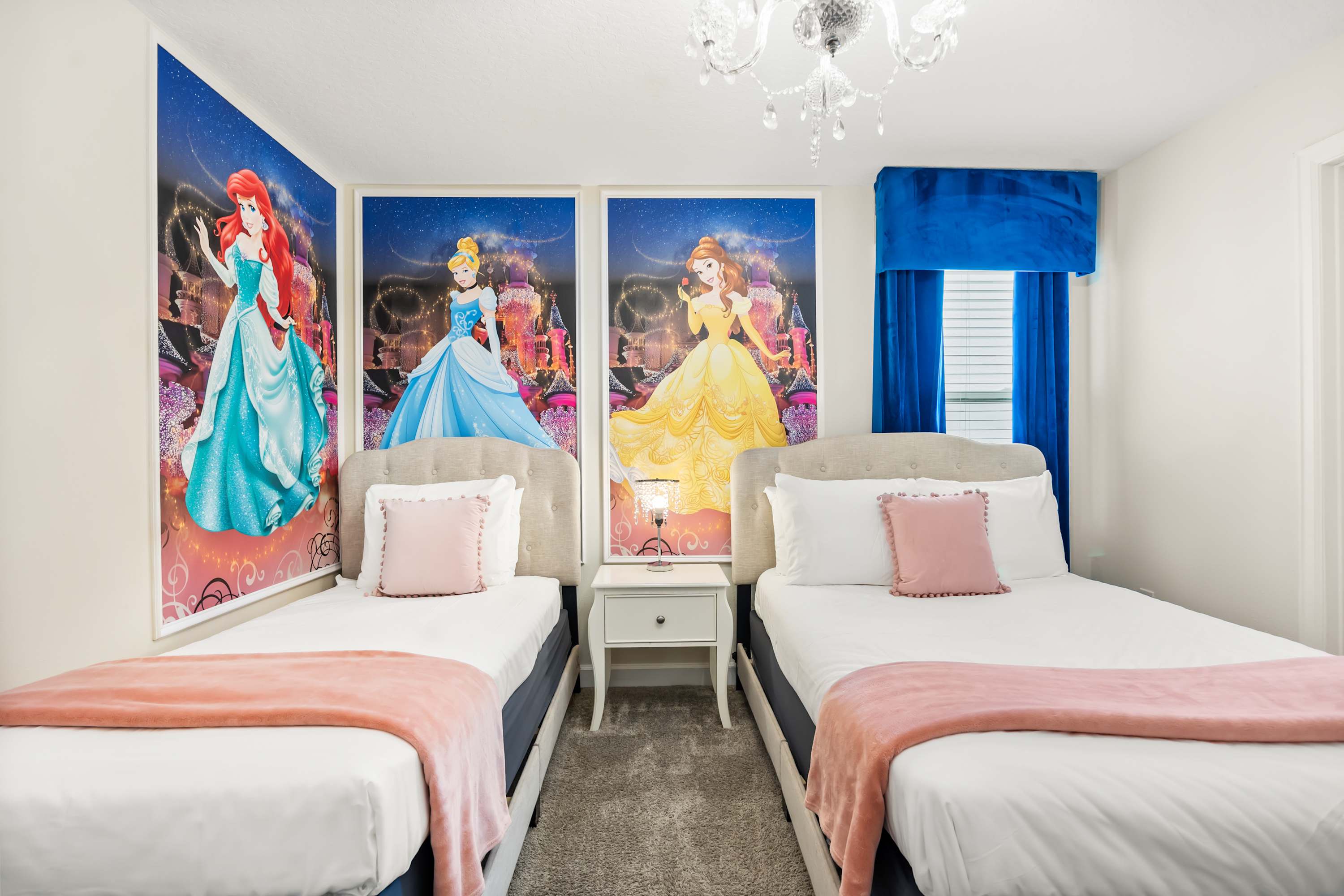 A vibrant kids' bedroom featuring two twin beds with pink throws and pillows, flanked by large wall murals of Disney princesses Ariel, Cinderella, and Belle, under a crystal chandelier.