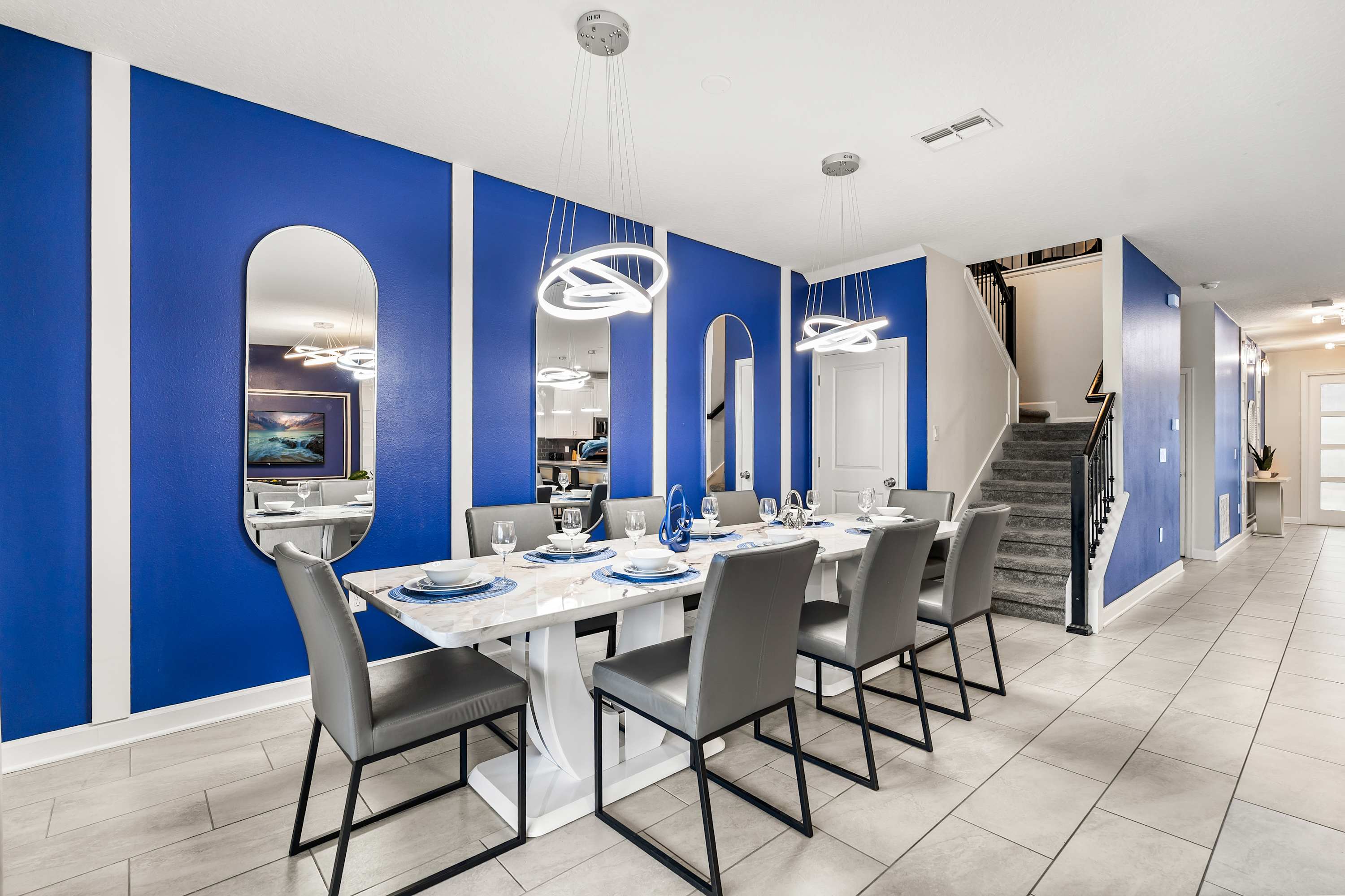 Contemporary dining room featuring a long white marble-top table with eight grey chairs, set against a vibrant blue accent wall with decorative mirrors and modern circular chandeliers.