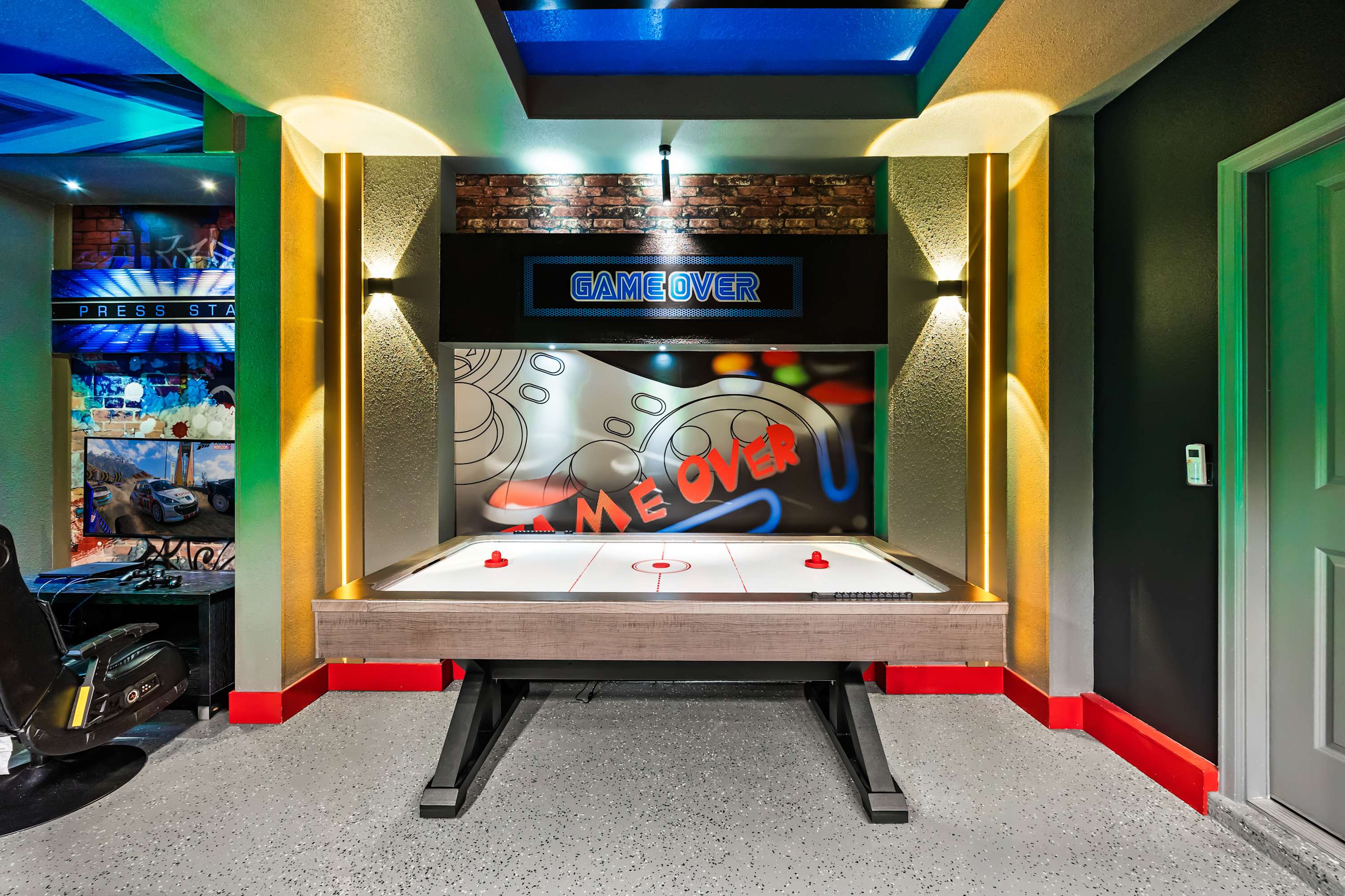 A vibrant game room features an air hockey table with red paddles, illuminated by colorful LED strip lighting on the walls and ceiling. A "GAME OVER" sign glows above a large graphic display, creating an energetic atmosphere.