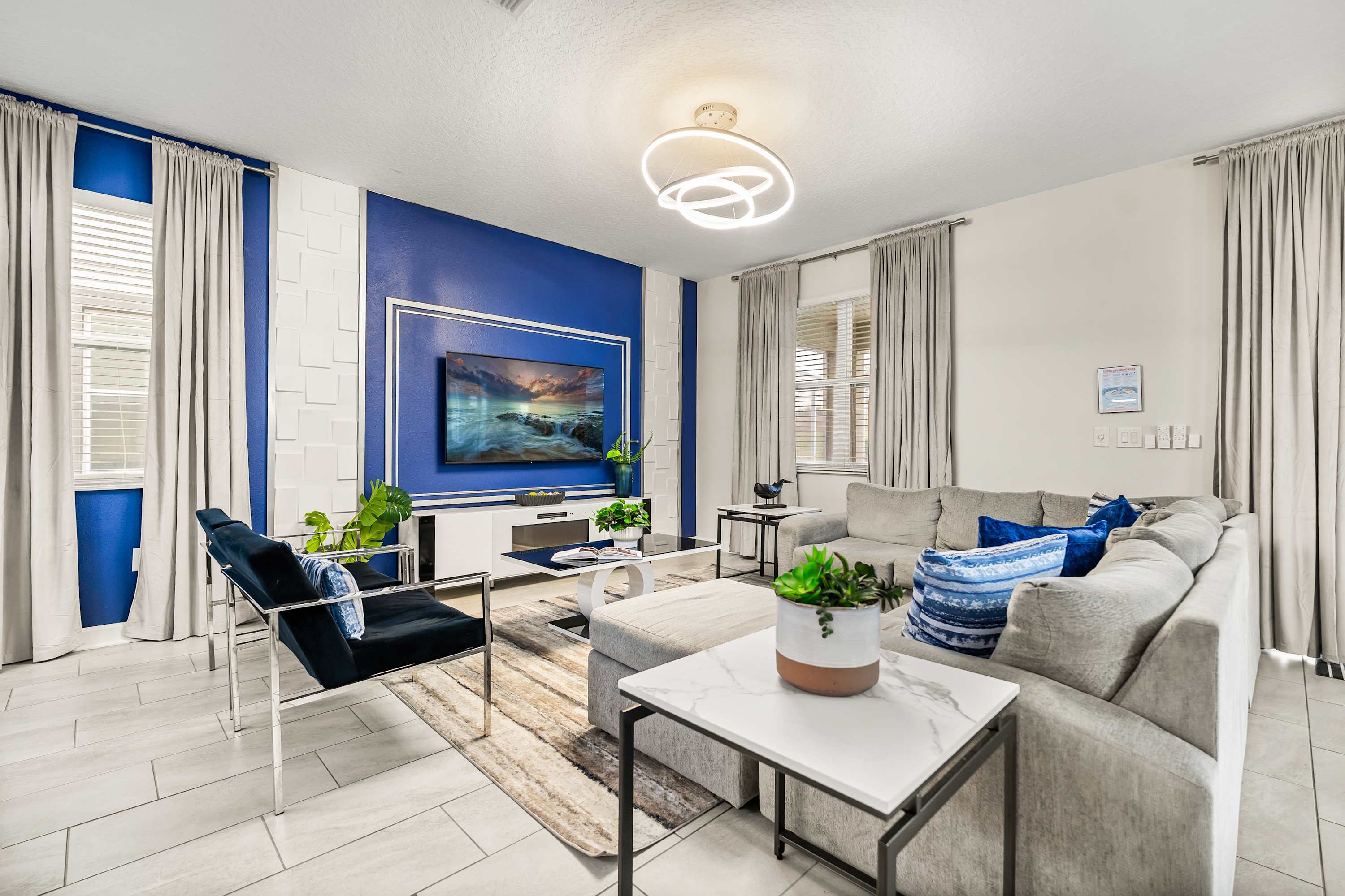 A contemporary living room showcases a large gray sectional sofa with blue pillows, facing a wall-mounted TV above a white console. The accent wall features a striking blue panel framed by white geometric 3D tiles, complemented by sheer gray curtains and a modern circular chandelier.