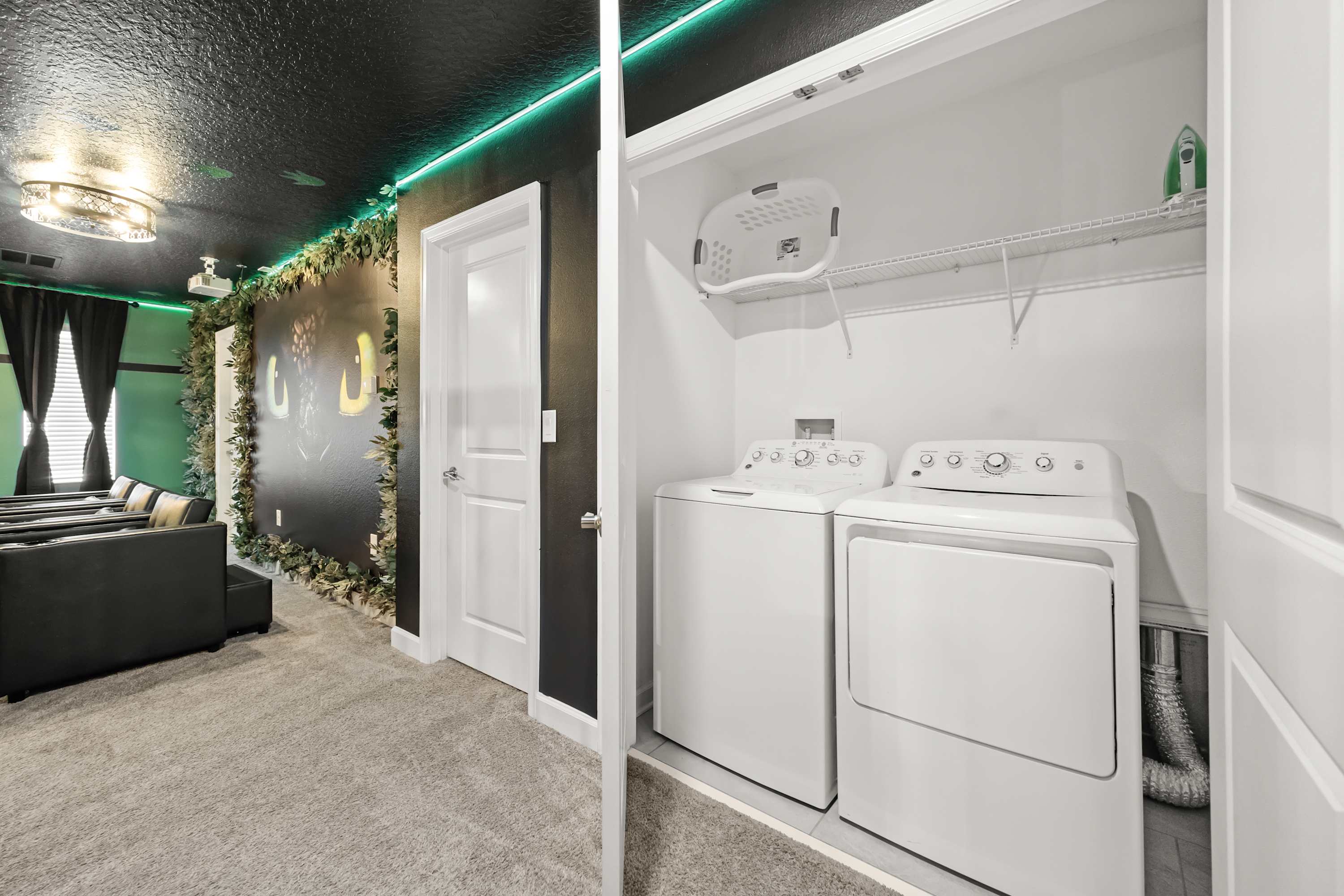 Laundry closet with a white top-loading washing machine and dryer, neatly tucked away. Adjacent is a room with a dark ceiling, green accent lighting, and a wall decorated with foliage, suggesting a themed entertainment space.