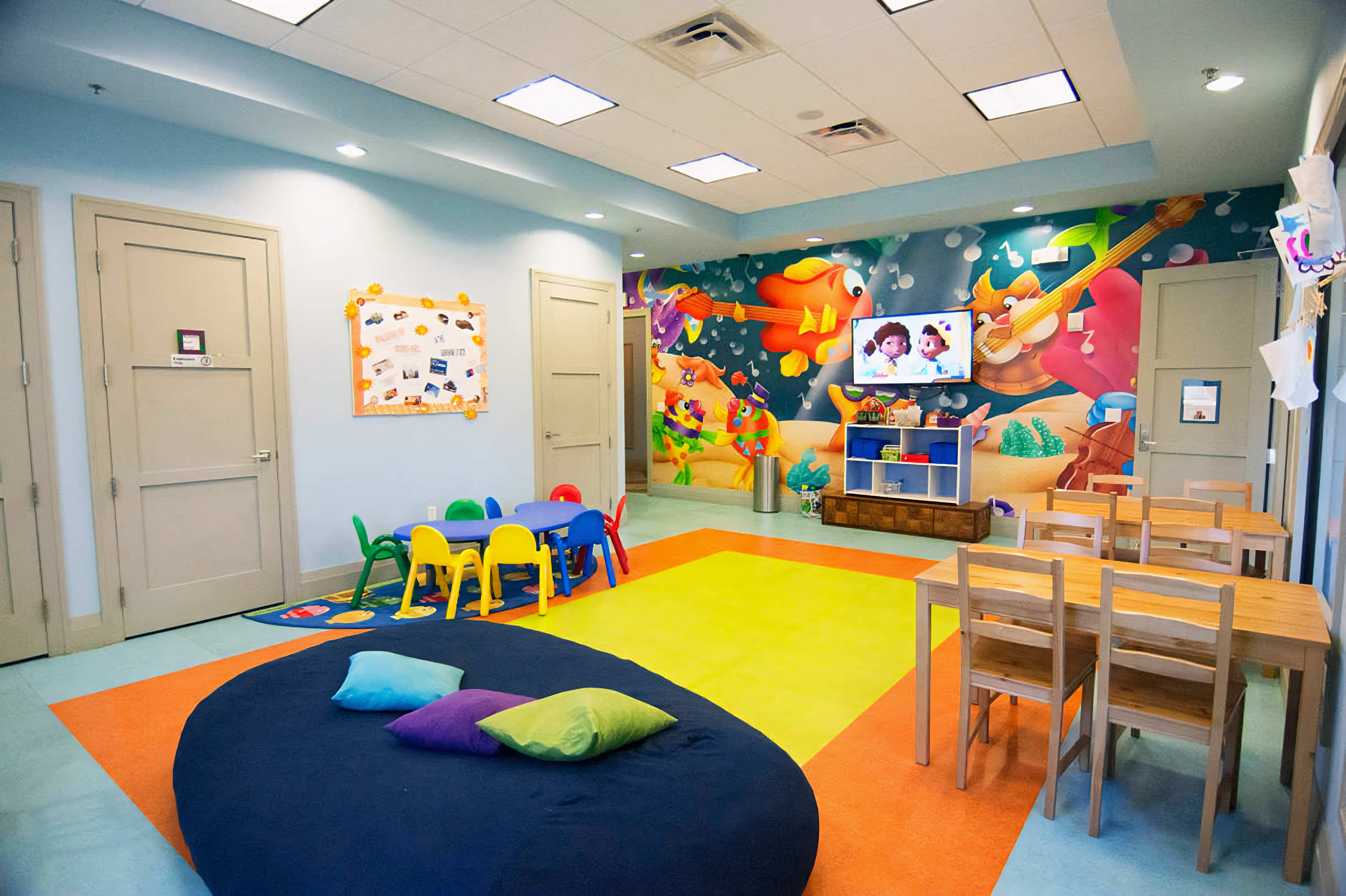 Colorful kids' club room with vibrant murals depicting musical instruments, featuring small tables and chairs, a large beanbag, and a TV playing cartoons, designed for children's activities.