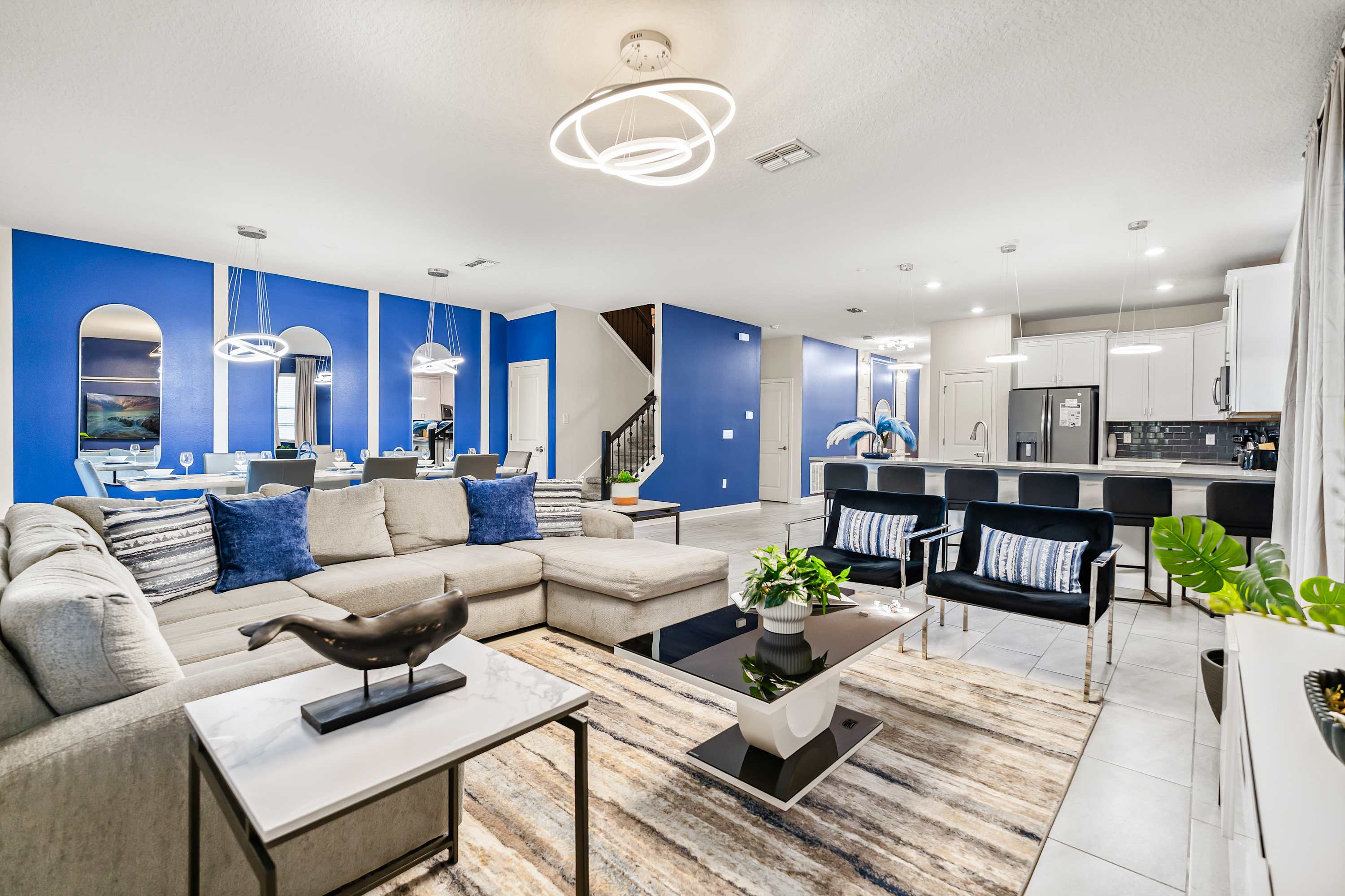 A spacious open-concept living room features a large L-shaped sectional sofa with blue throw pillows, facing a modern coffee table on a striped rug. Bold blue accent walls and contemporary circular light fixtures define the space, which opens to a dining area and kitchen.