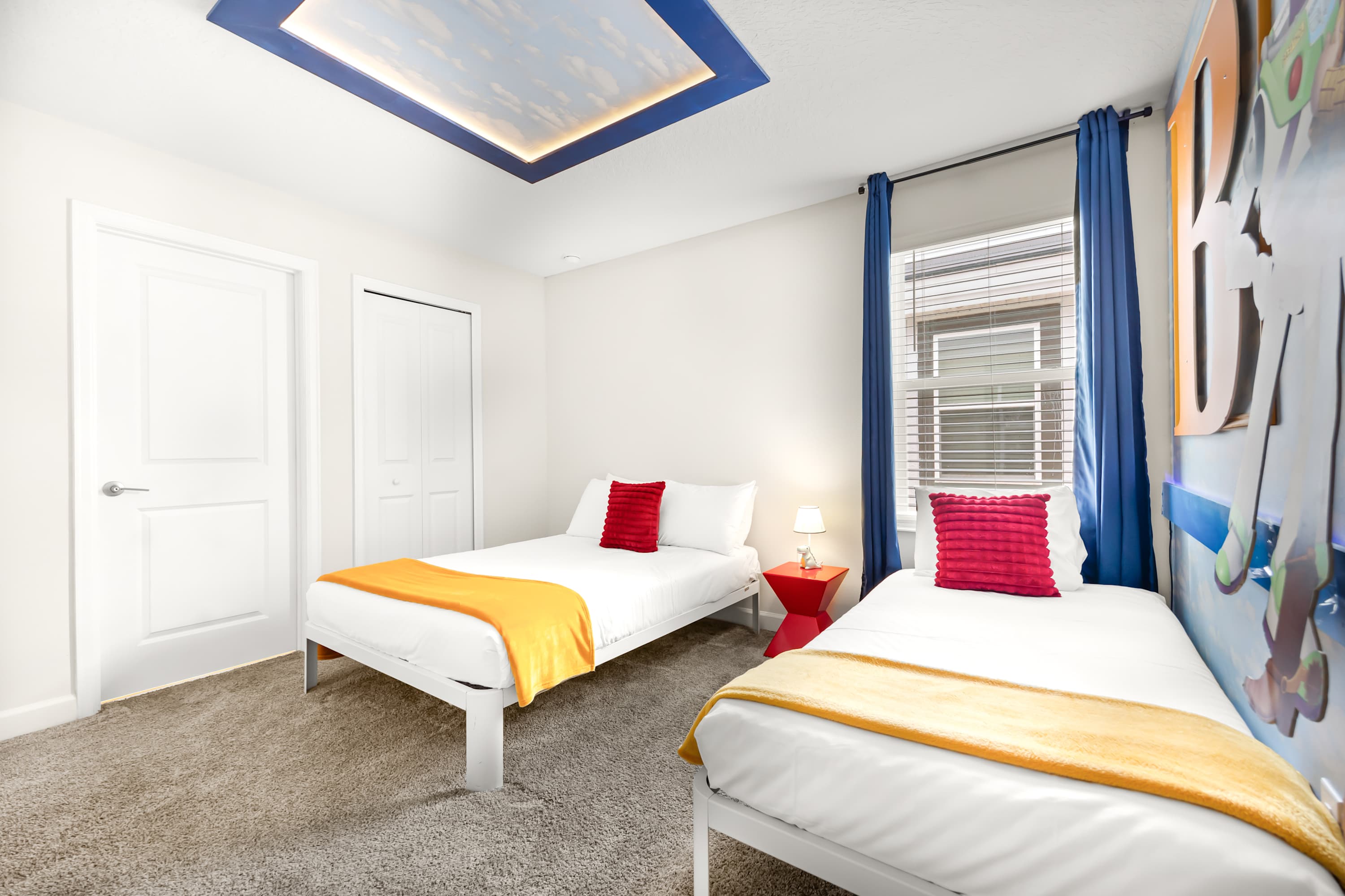 Another angle of the "Toy Story" themed bedroom, showing two single beds with red pillows and yellow throws, a red nightstand, and a window with blue curtains. White doors lead to a closet and another room.