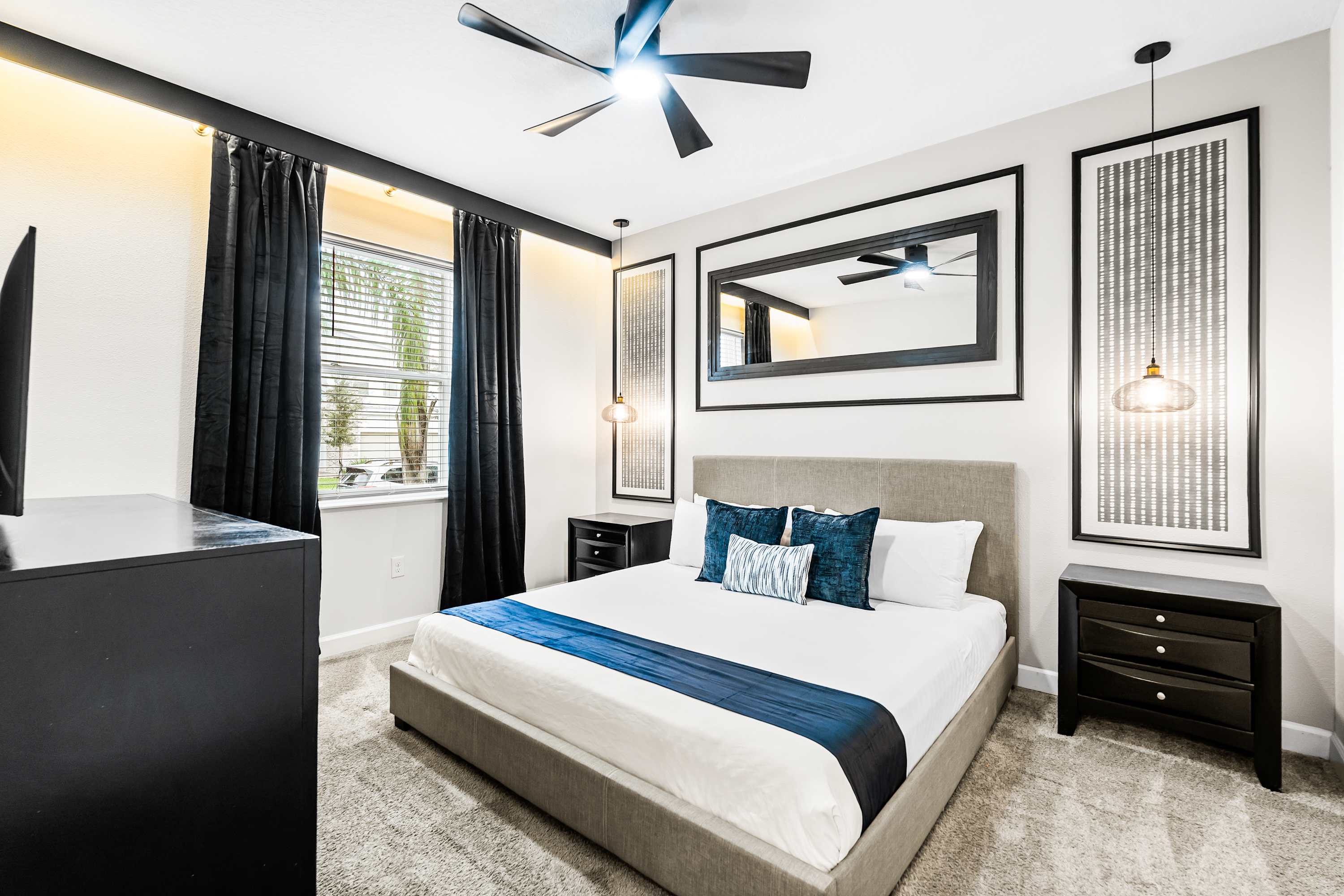 Modern bedroom featuring a gray upholstered king bed with white bedding and a blue runner. Black nightstands flank the bed, topped with pendant lights, and a large mirror with a black frame hangs above the headboard. Black curtains frame a window with blinds.