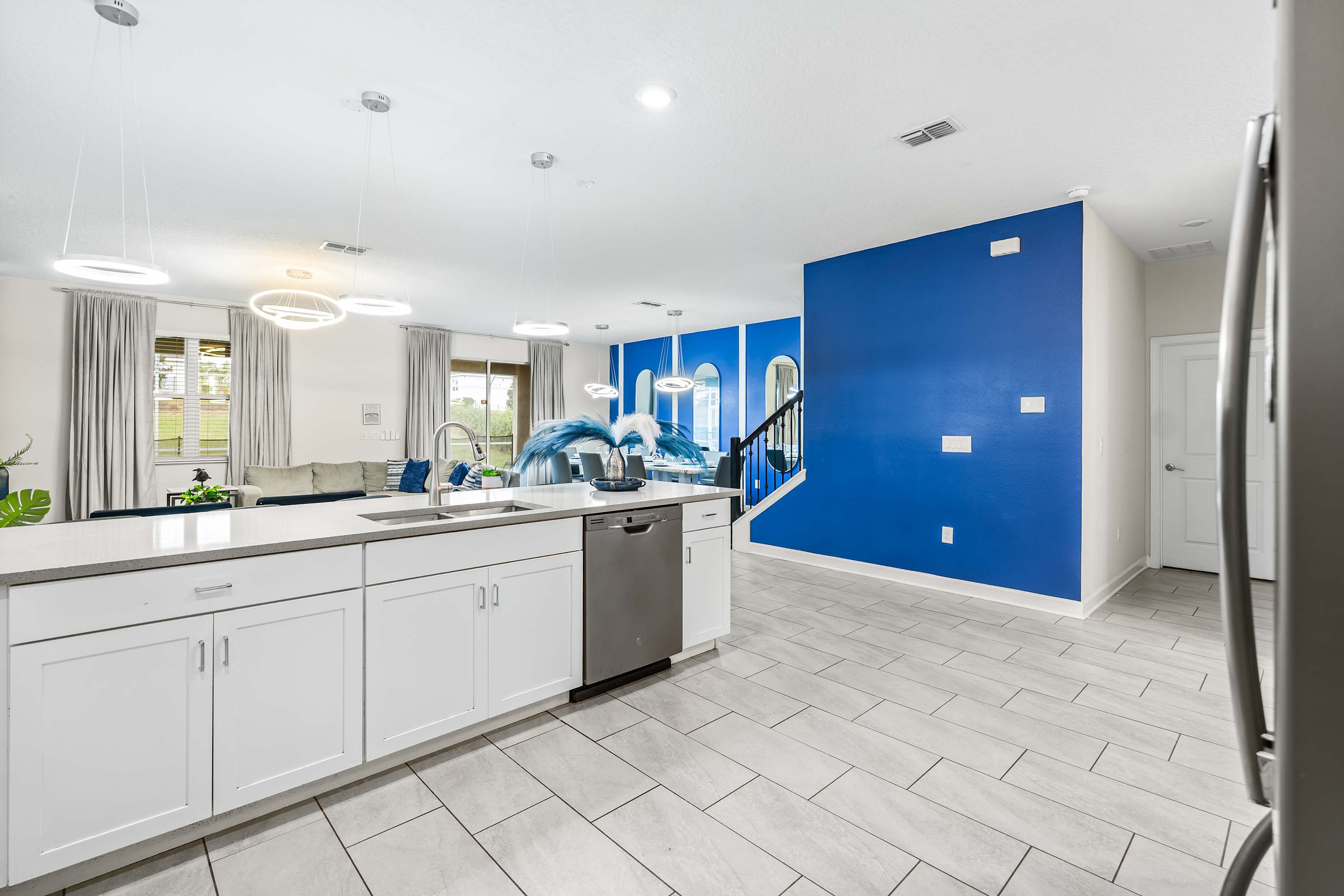 A view from the kitchen island looking towards the open living and dining areas, showcasing the seamless flow of the space. The kitchen features white cabinetry, a large island with a sink, and modern pendant lighting, with a vibrant blue accent wall visible in the background.