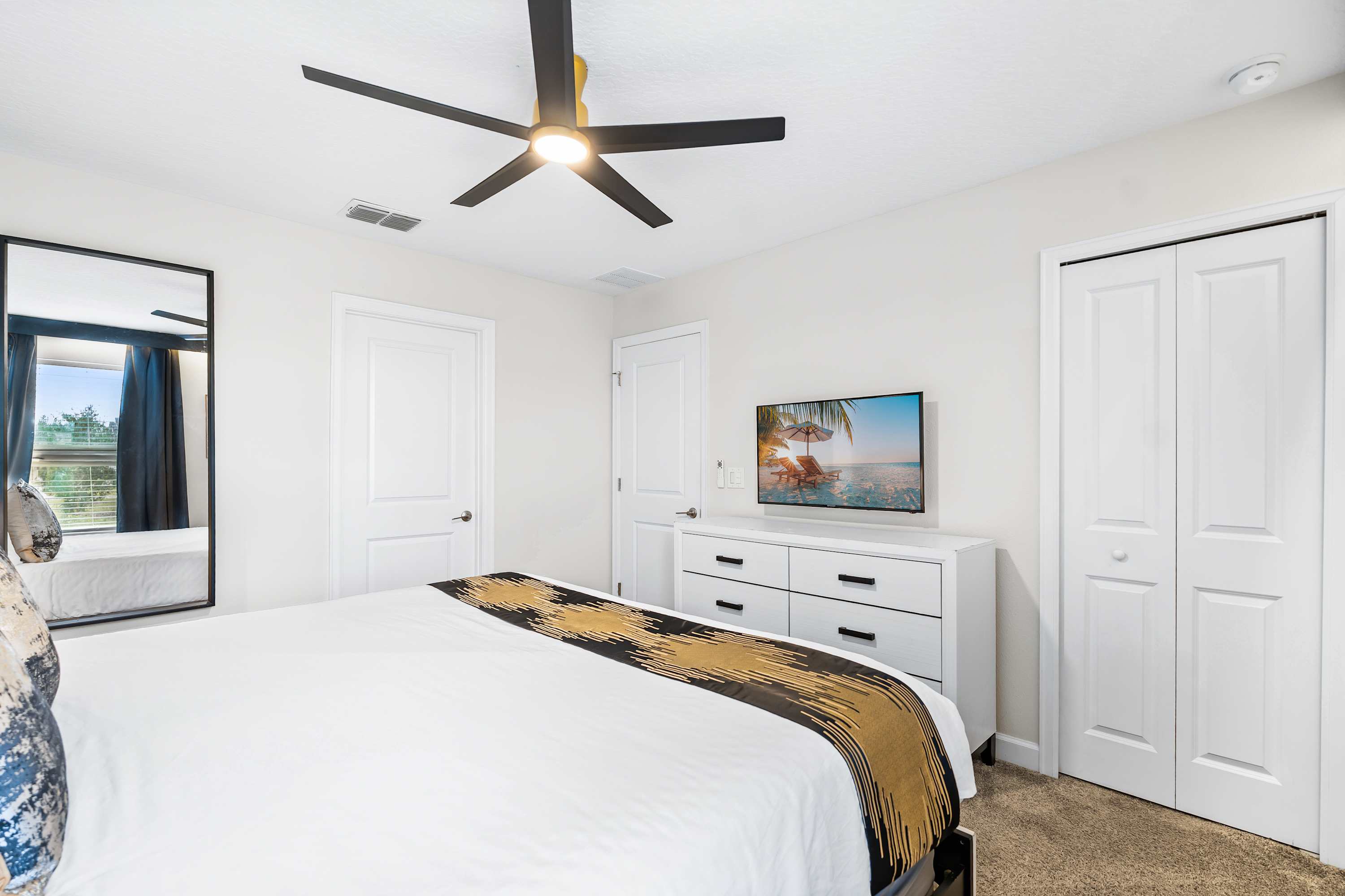 A different perspective of the stylish bedroom, showing a white dresser with a flat-screen TV, a full-length mirror, and white closet doors, complementing the room's modern design.
