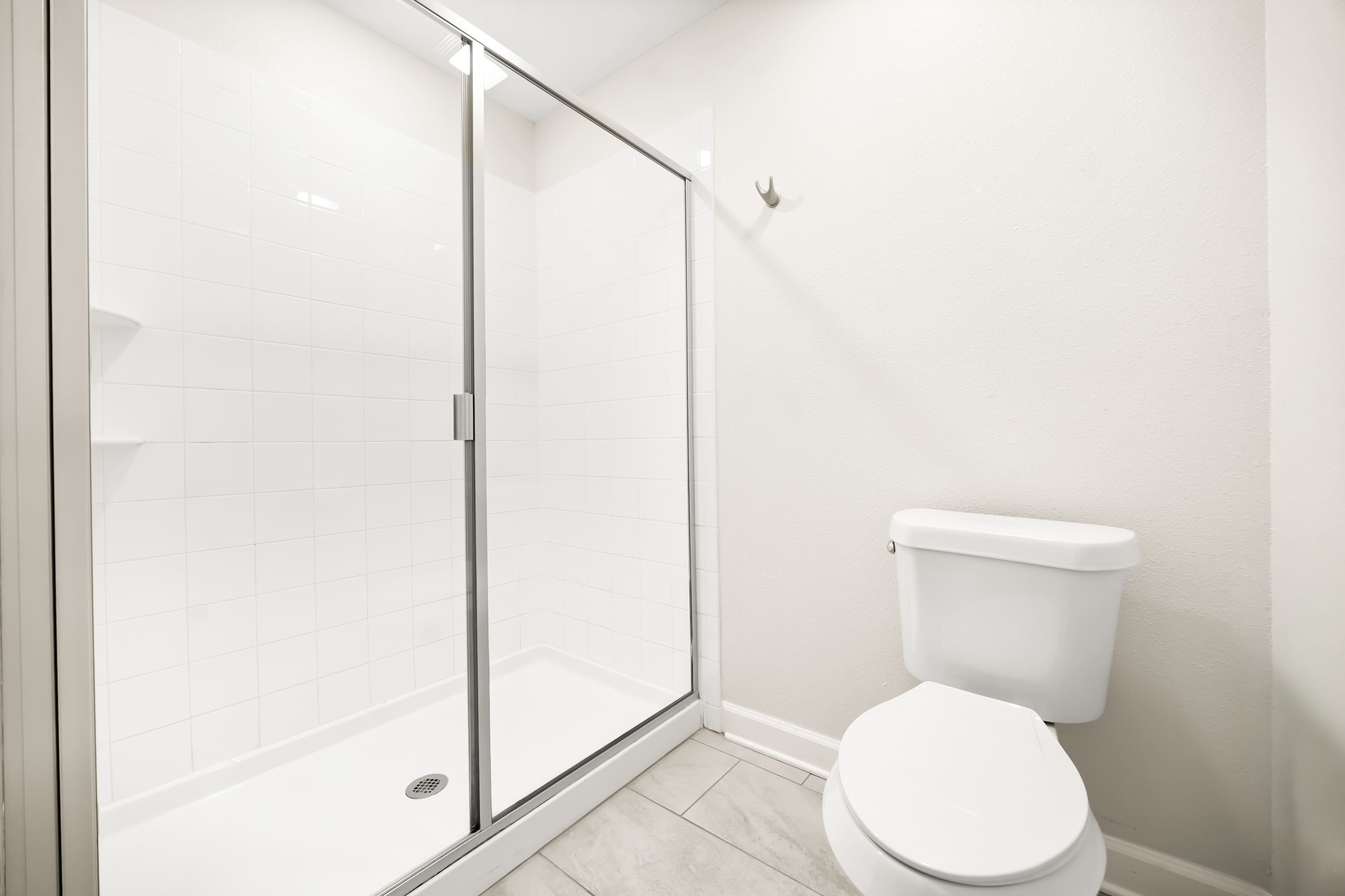 A clean, modern bathroom section with a white toilet and a glass-enclosed shower featuring white subway tiles. Light gray tiled floors match the adjacent vanity area.