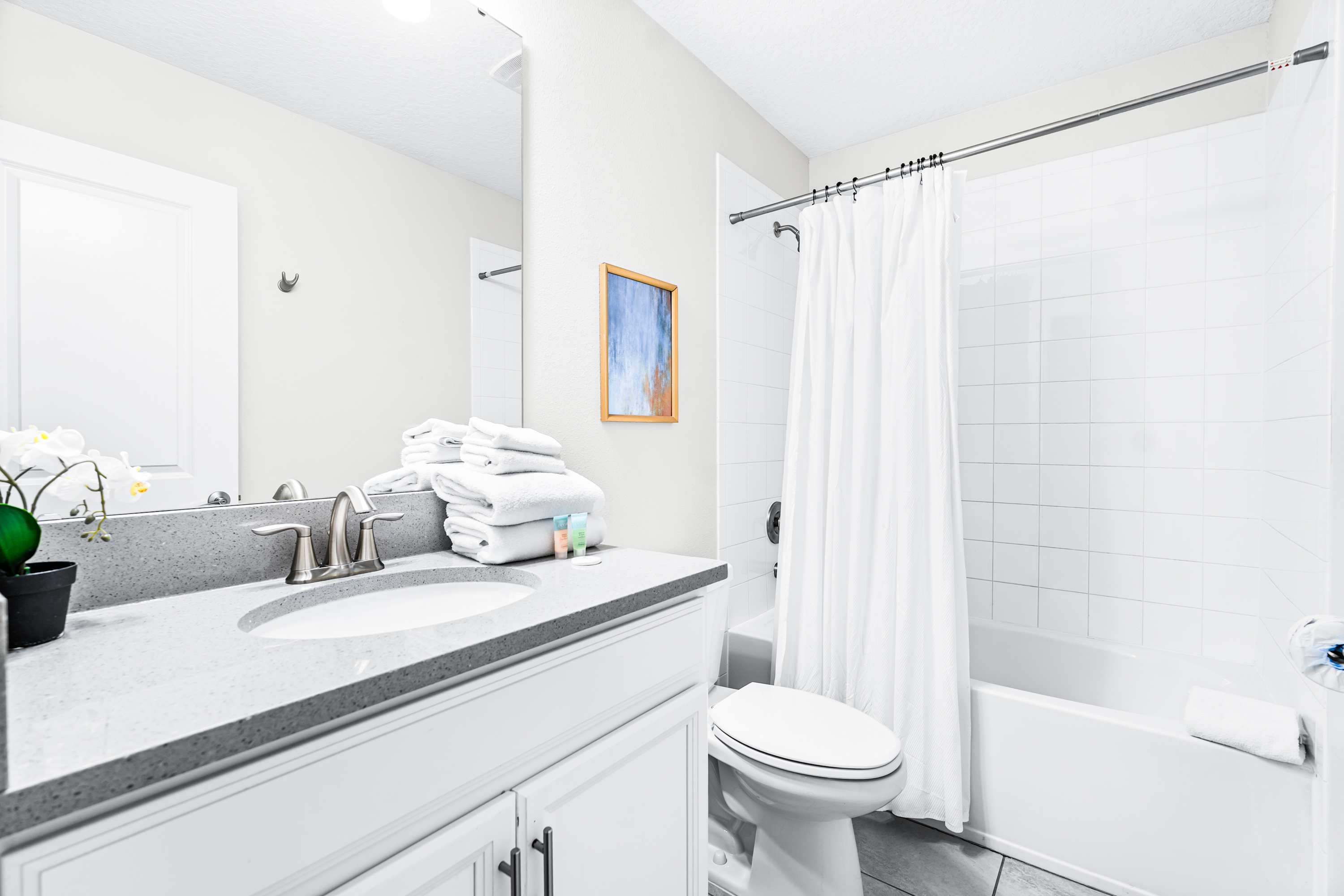 A clean, white bathroom featuring a single vanity with a light gray countertop, a large mirror, and a bathtub with a shower curtain, stocked with fresh towels and toiletries.