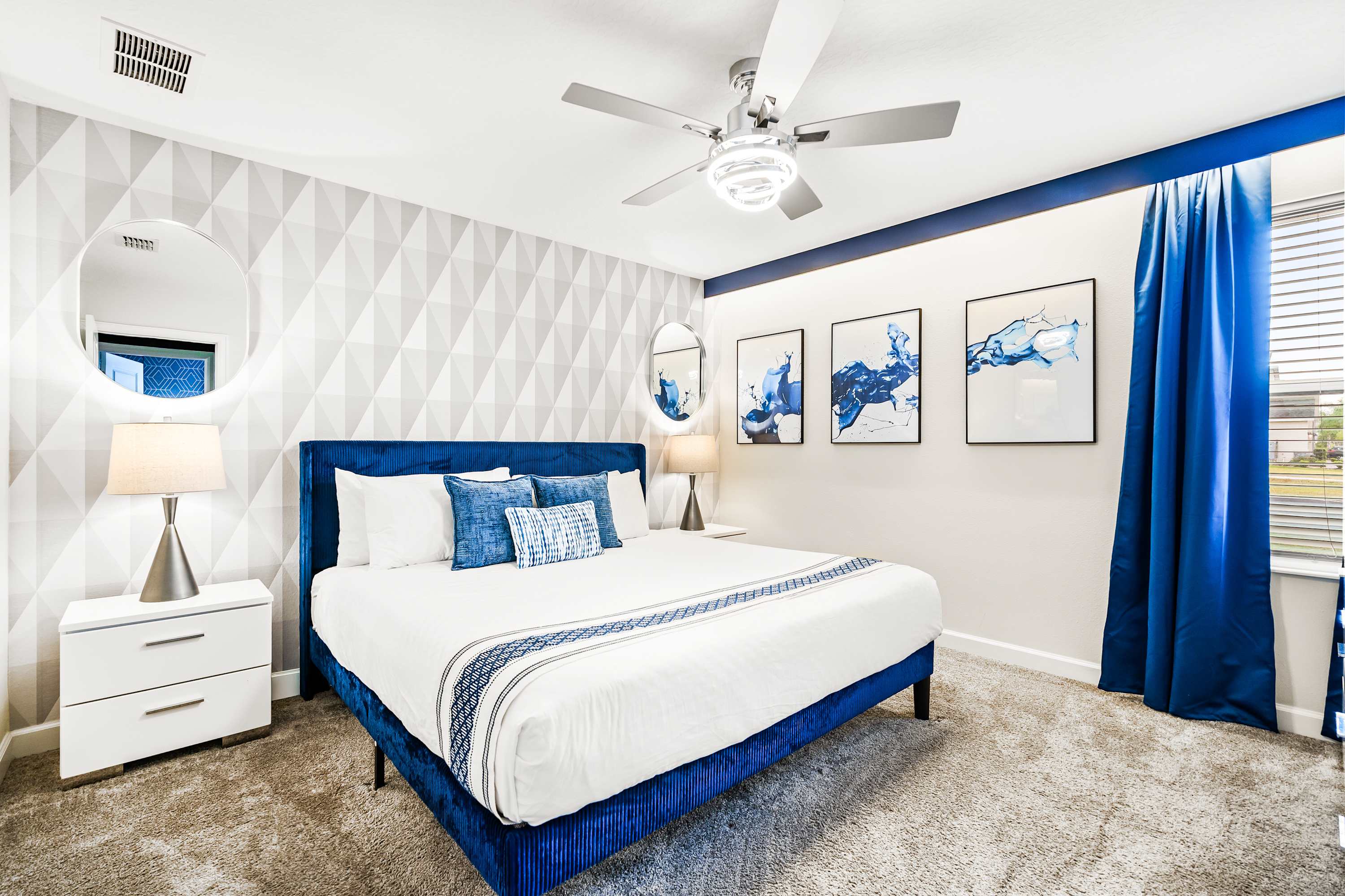 A modern bedroom featuring a king-sized bed with a blue velvet headboard, white bedding, and blue accent pillows, complemented by geometric wallpaper and blue curtains.