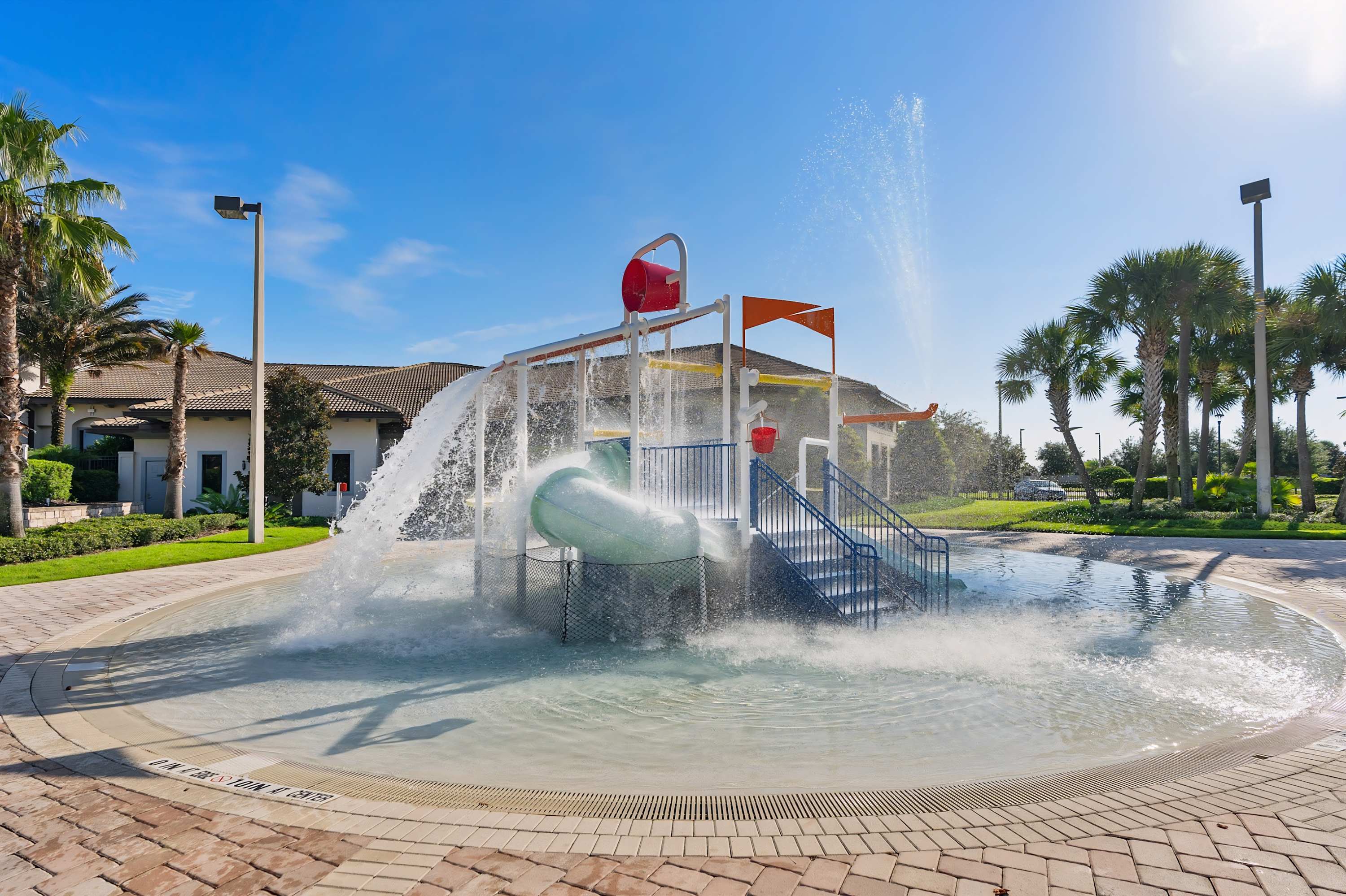 Vibrant splash pad area designed for children, featuring a large water play structure with buckets, slides, and spraying fountains, set on a patterned paved surface amidst palm trees.