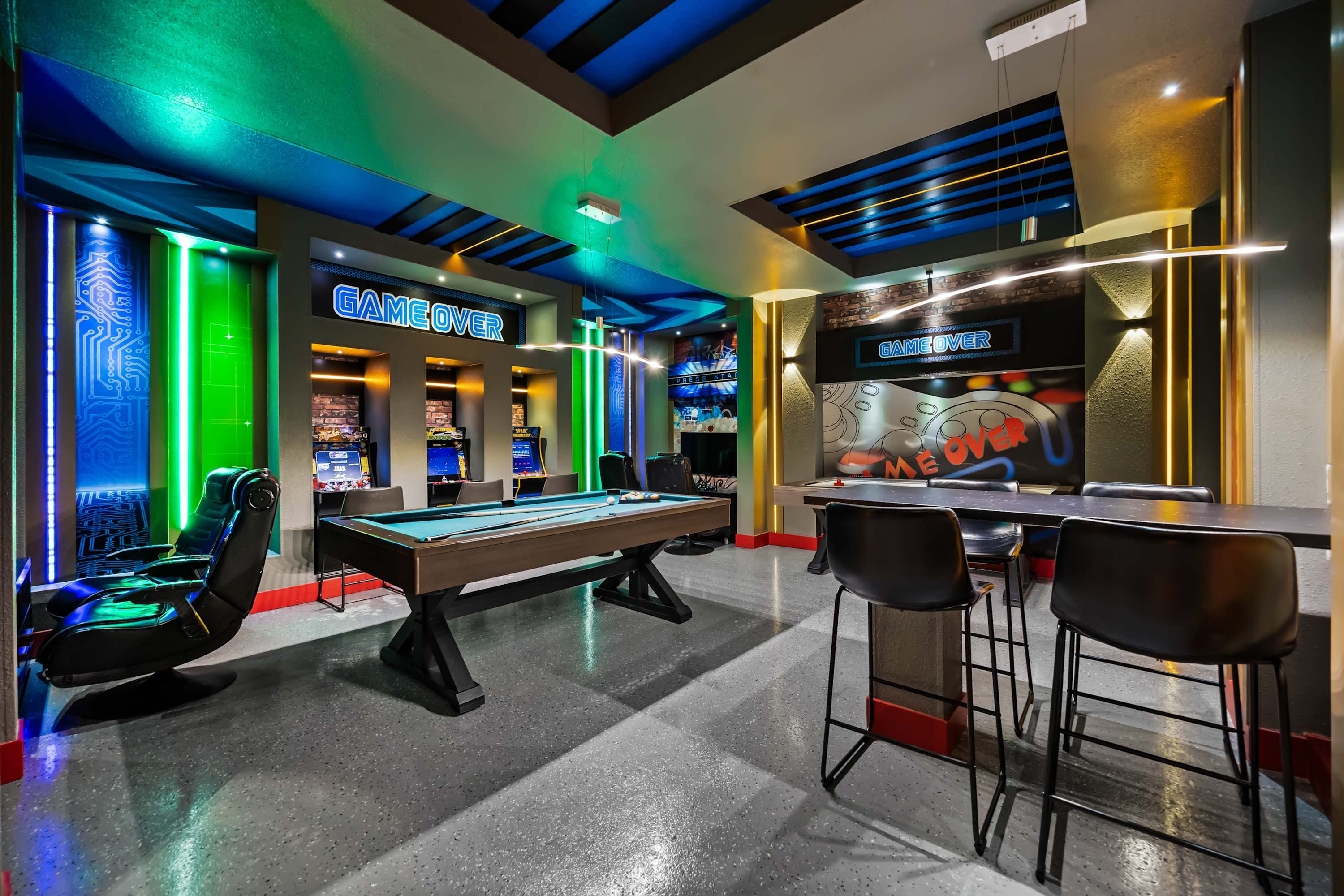 A vibrant game room features a pool table in the center, surrounded by arcade machines and a high-top table with stools. Neon green and blue LED lighting illuminates the walls, creating an energetic and playful atmosphere.