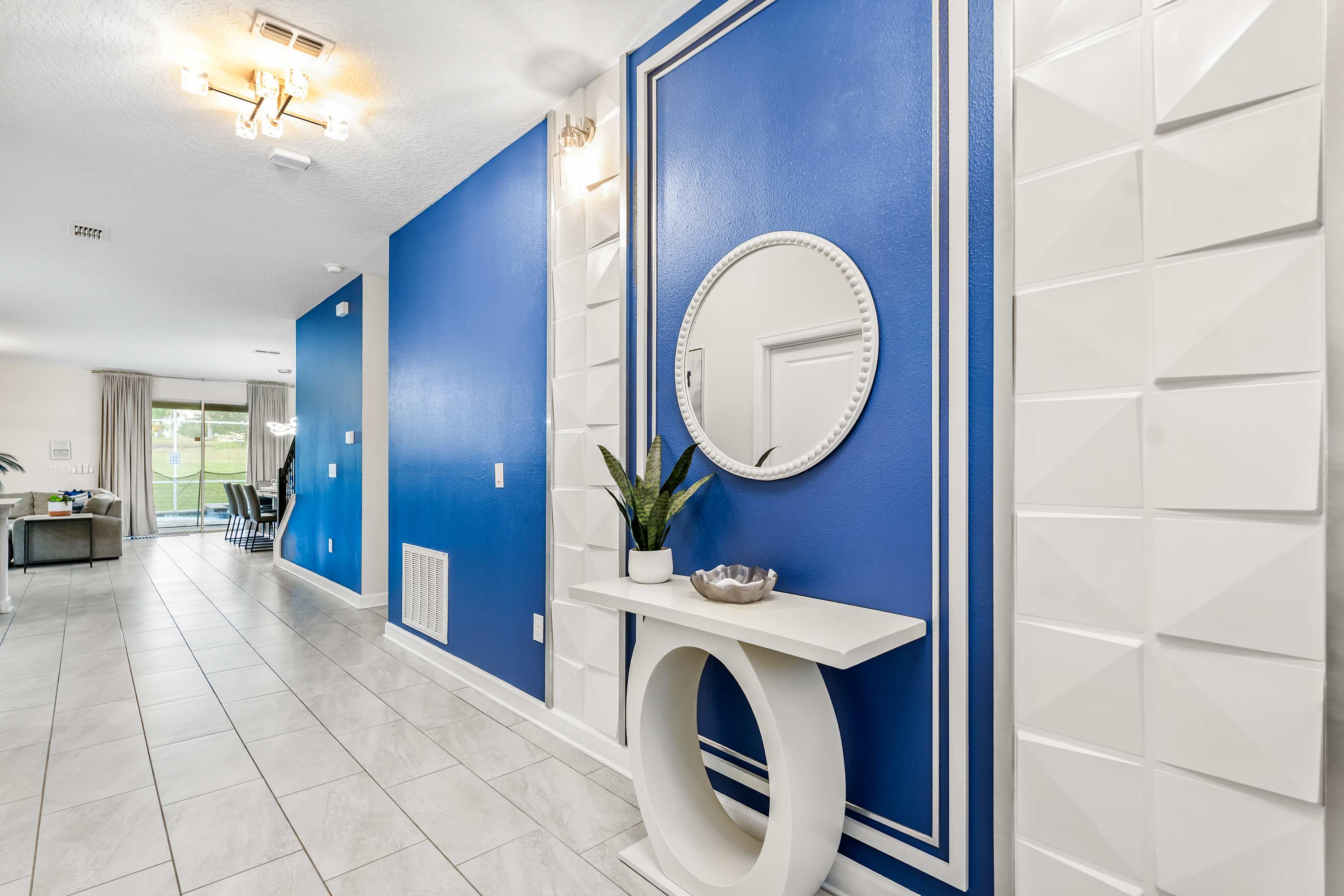 A bright foyer features a vibrant blue accent wall adorned with white geometric 3D panels and a round mirror above a white console table. The space is illuminated by a modern ceiling light and leads into the open-concept living areas.
