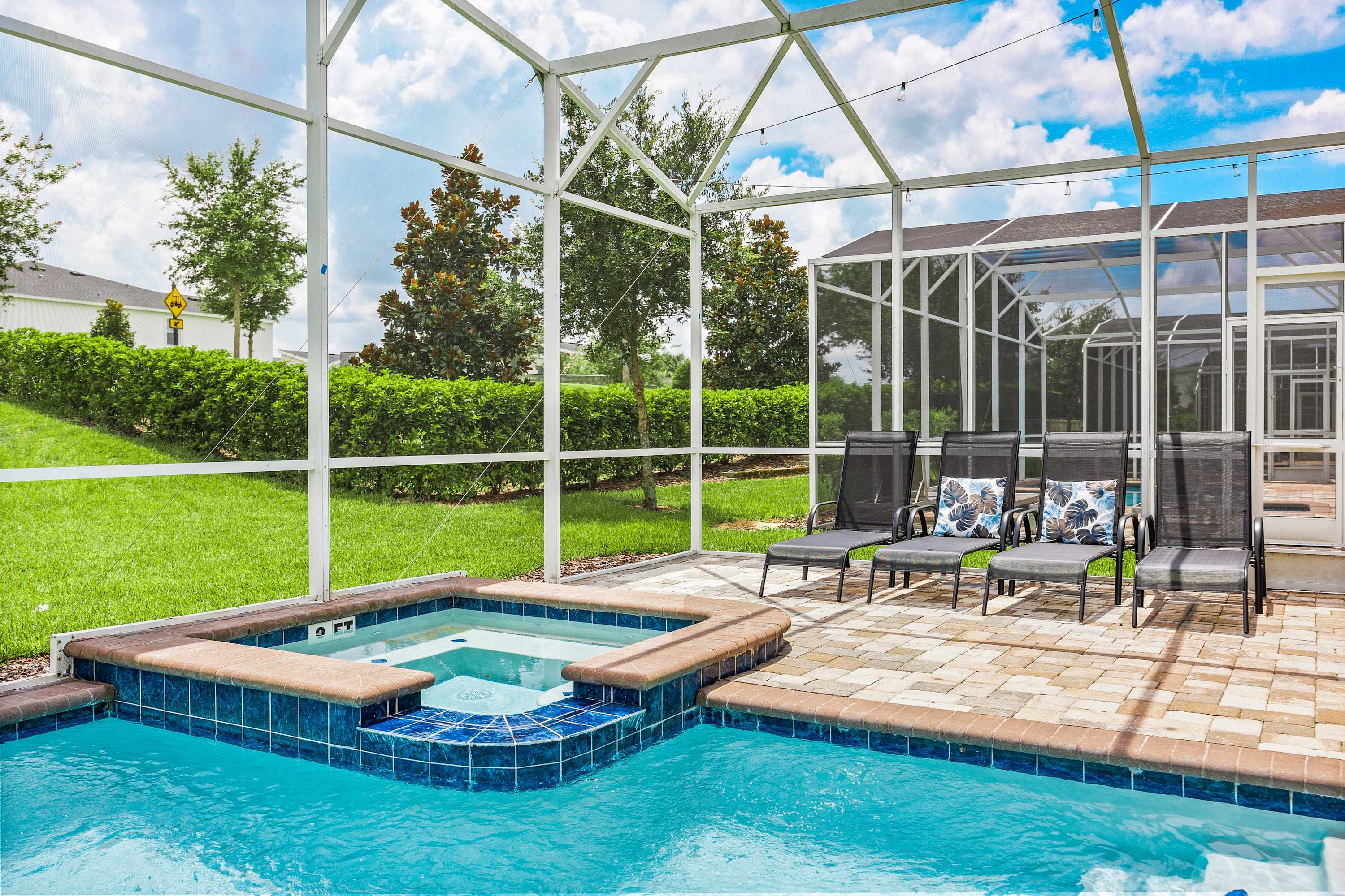 Close-up of a square hot tub with blue tile accents, surrounded by a paver deck with several dark lounge chairs and decorative pillows, all within a large screened enclosure.