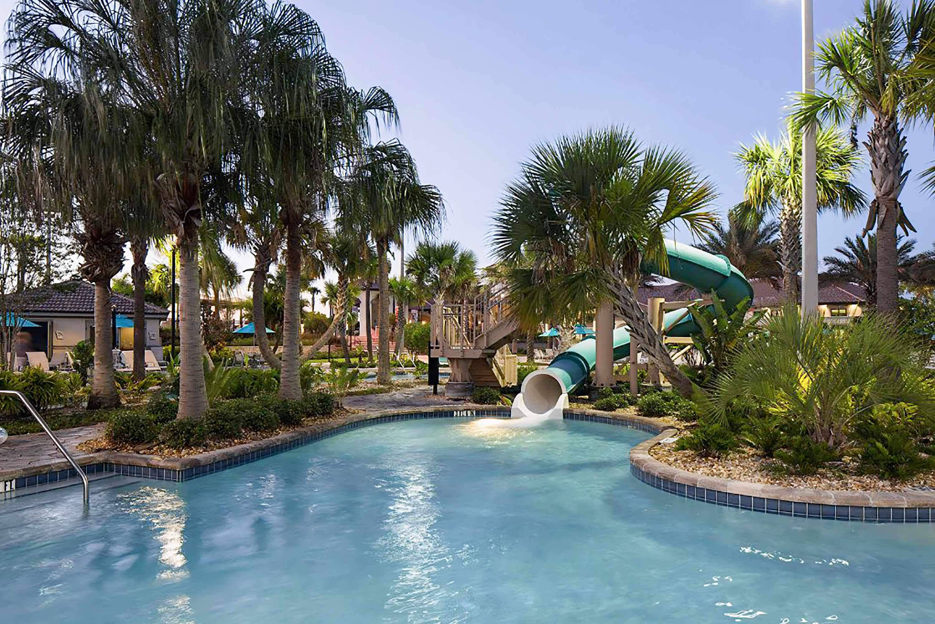 Lush communal pool area featuring a winding water slide that empties into a shallow pool, surrounded by numerous palm trees and tropical foliage under a bright sky.