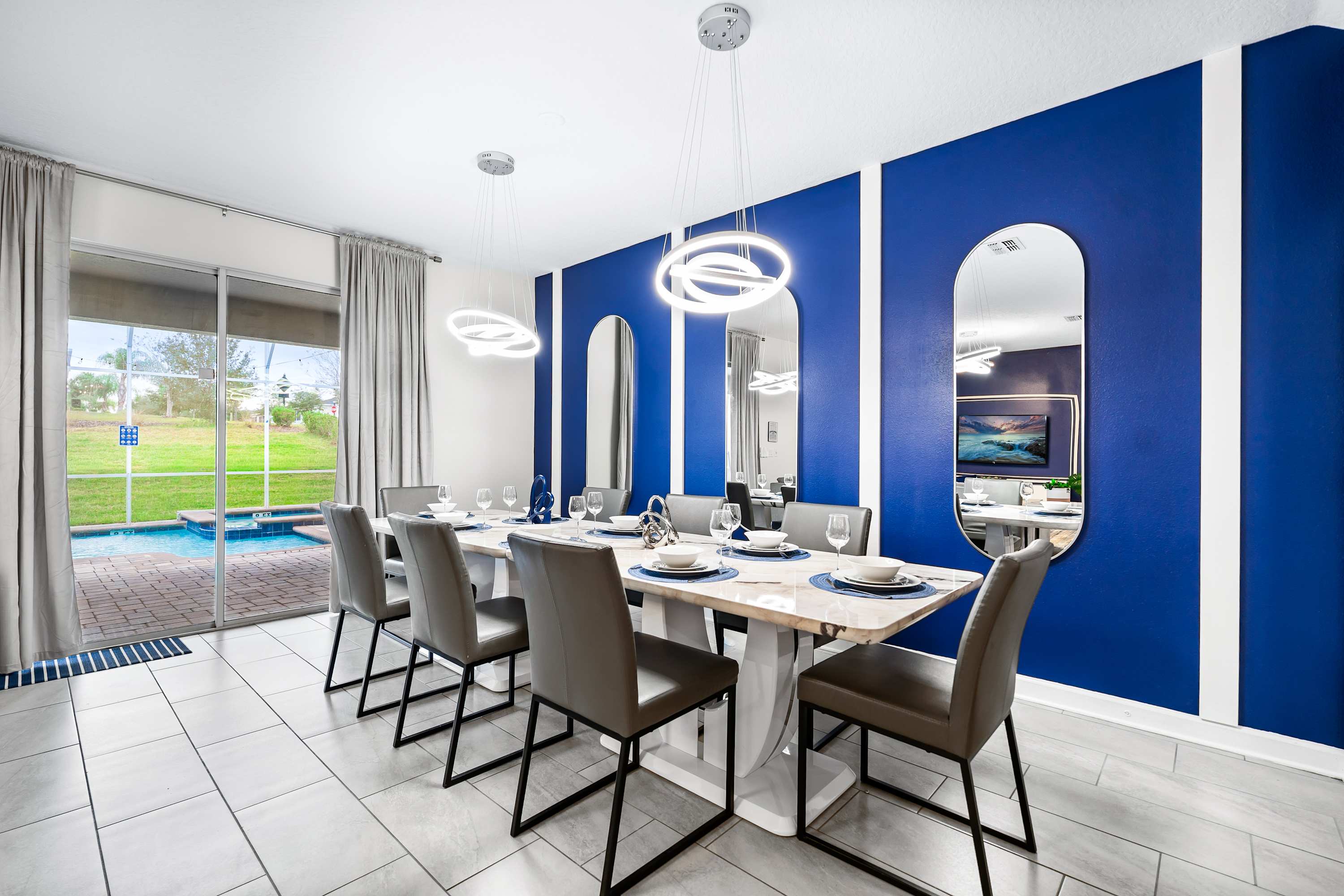 A stylish dining room features a long white marble-top table with seating for eight, set against a vibrant blue accent wall with three tall arched mirrors. Modern circular pendant lights hang above, and sliding glass doors lead to an outdoor pool area.