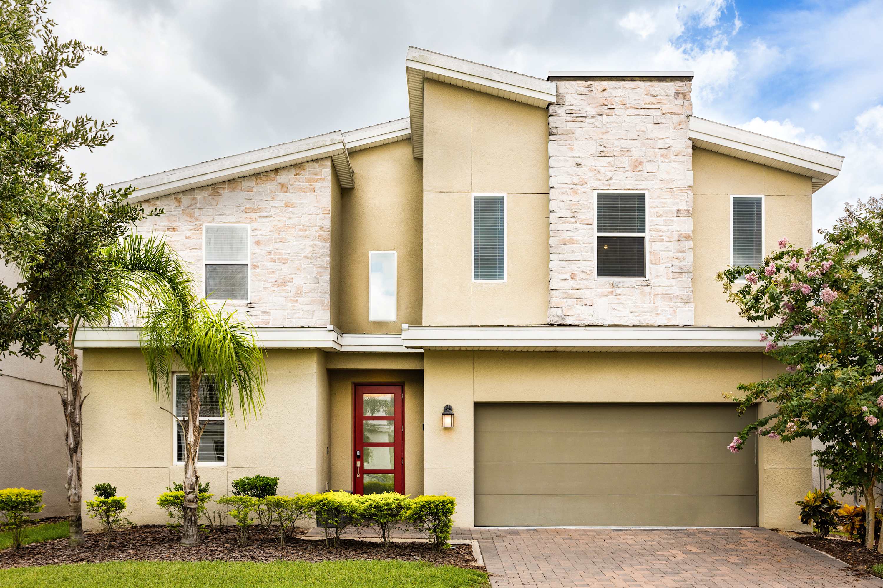 Modern two-story house with a light beige stucco exterior and stone accents. It features a red front door, a two-car garage with a brown door, and a paved driveway, surrounded by green landscaping and palm trees.