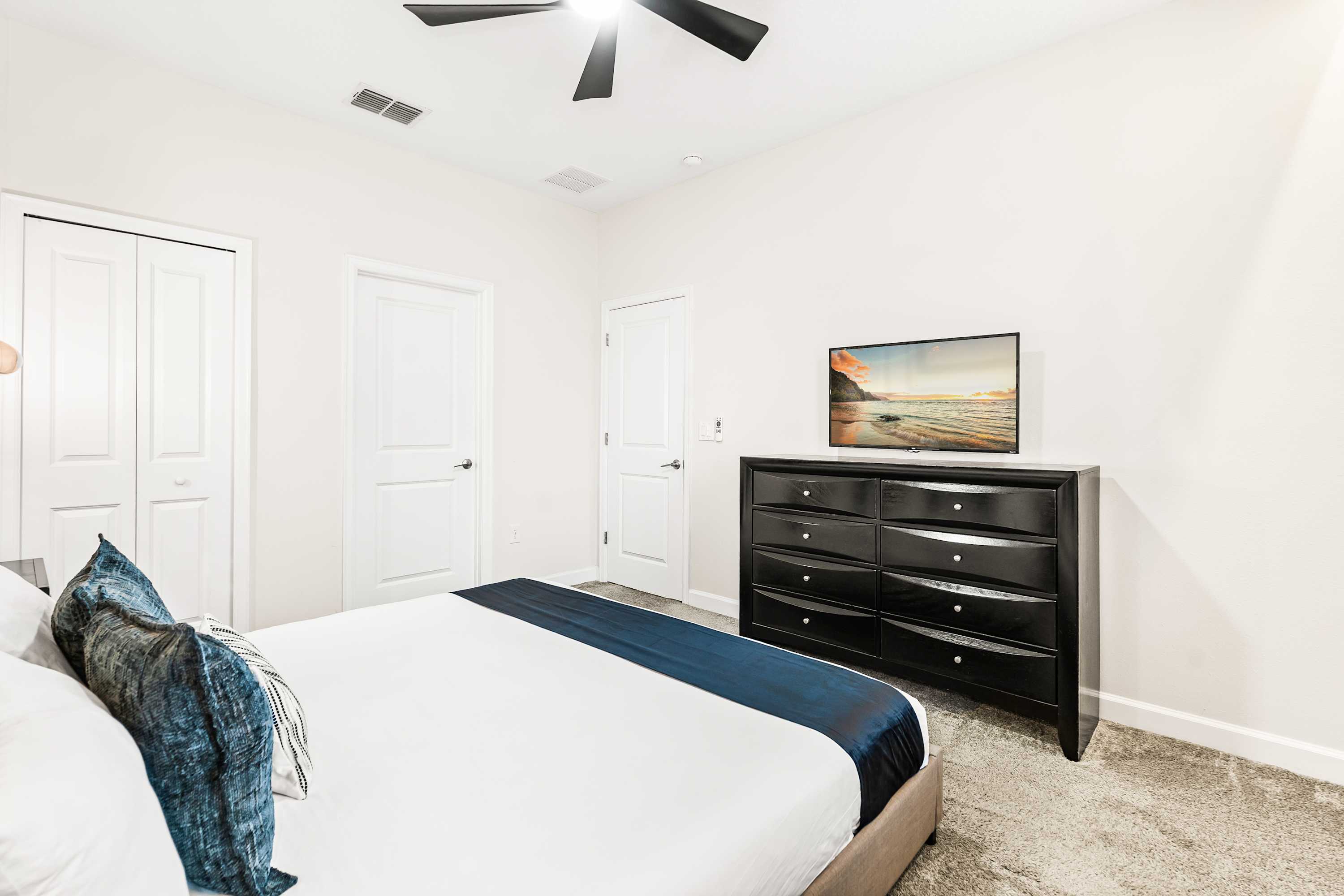 Bright bedroom featuring a large black dresser with a flat-screen TV on top, set against a white wall. A white bed with blue accents is partially visible in the foreground, and two white doors are seen in the background.