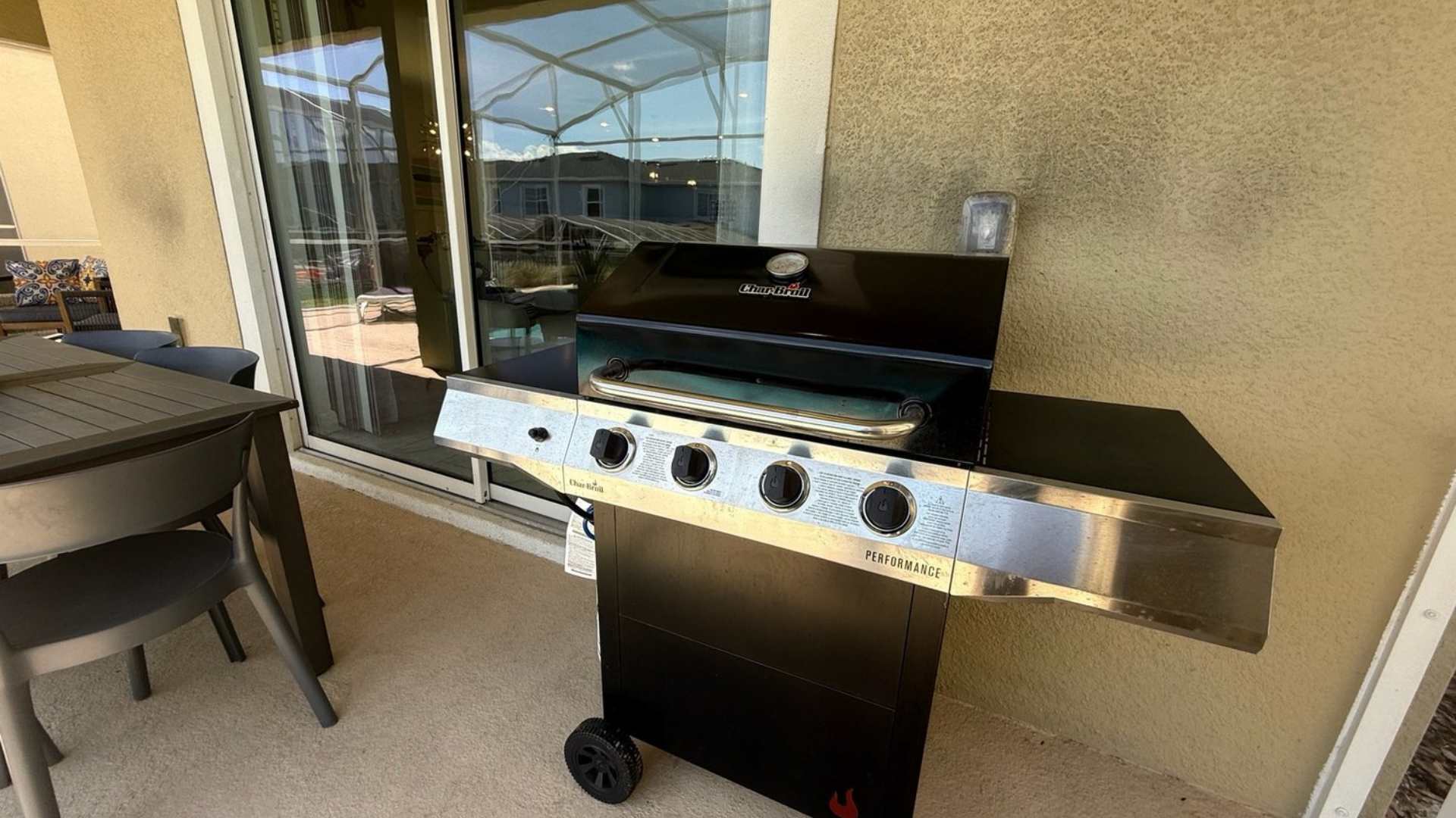Outdoor patio area featuring a black Char-Broil gas grill with stainless steel accents, positioned next to an outdoor dining table, with sliding glass doors leading indoors.