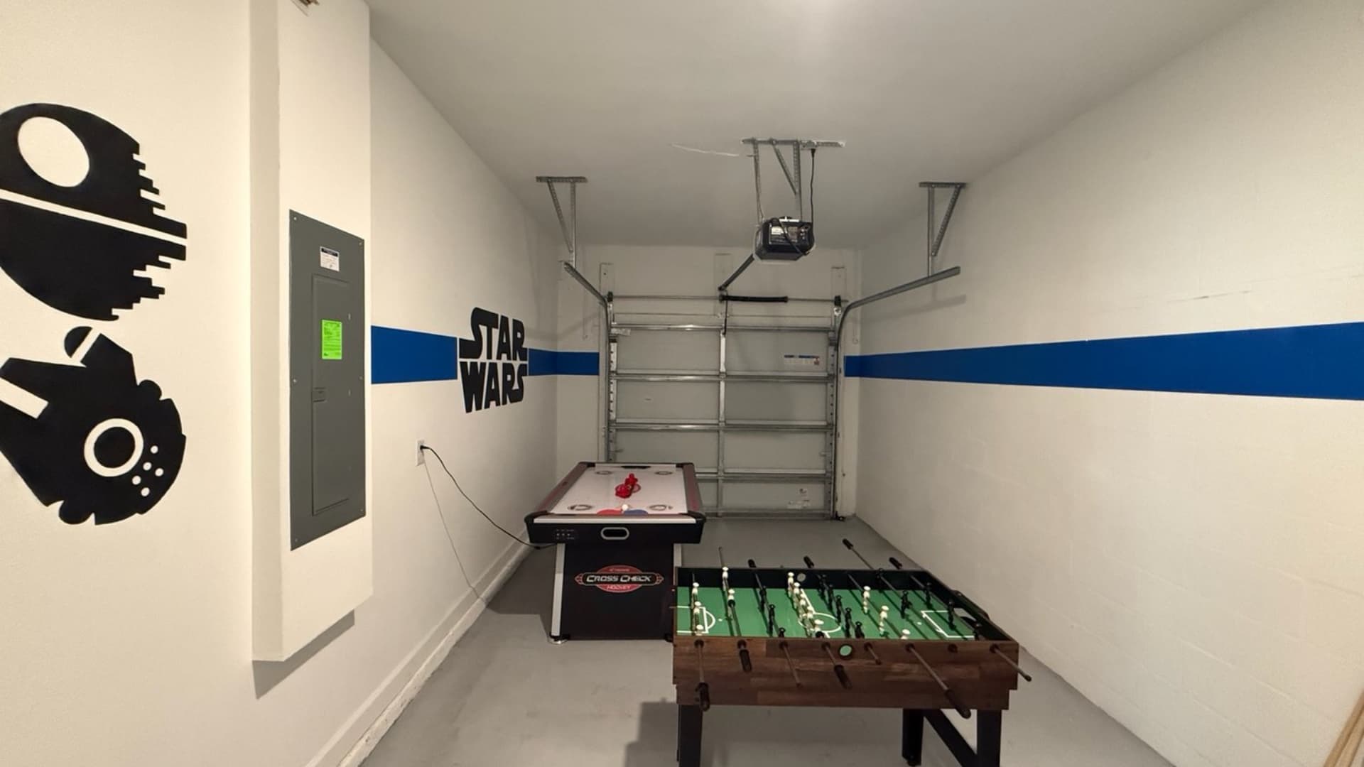 Garage converted into a game room featuring an air hockey table and a foosball table. The white walls are decorated with Star Wars themed decals and a blue stripe.