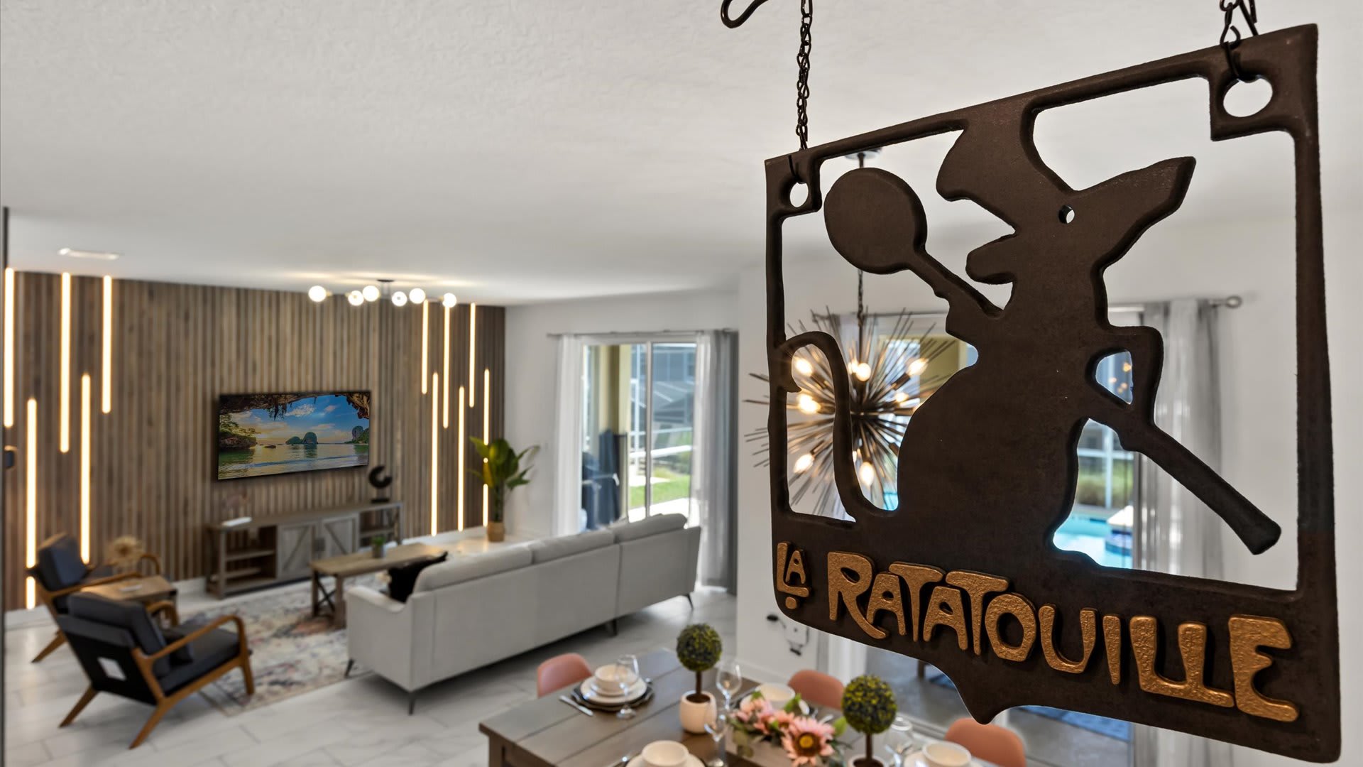 An elevated view captures the open-concept living and dining areas, featuring a "La Ratatouille" sign hanging in the foreground. The living room has a wooden slatted accent wall with LED lights and a TV, while the dining area boasts a modern chandelier and a wooden table with pink chairs.
