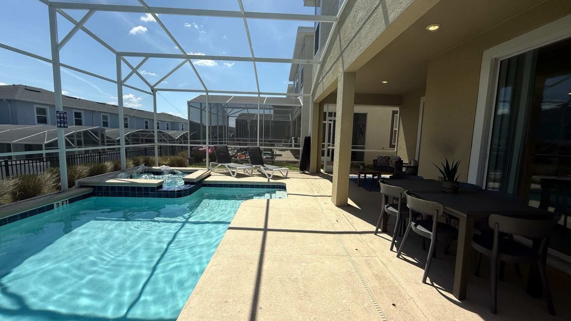 Expansive screened-in pool deck with a clear blue swimming pool and hot tub, offering views of neighboring houses, and a covered patio with outdoor dining furniture.