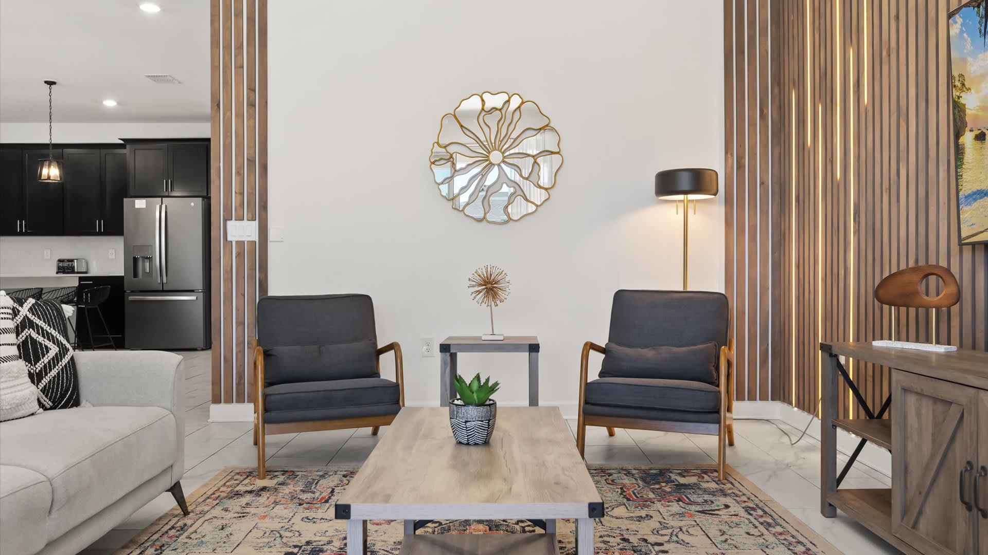 This living room view highlights two dark gray armchairs with wooden frames flanking a small side table, positioned on a colorful patterned rug. A large gold floral mirror adorns the white wall, and a tall black floor lamp provides additional lighting.