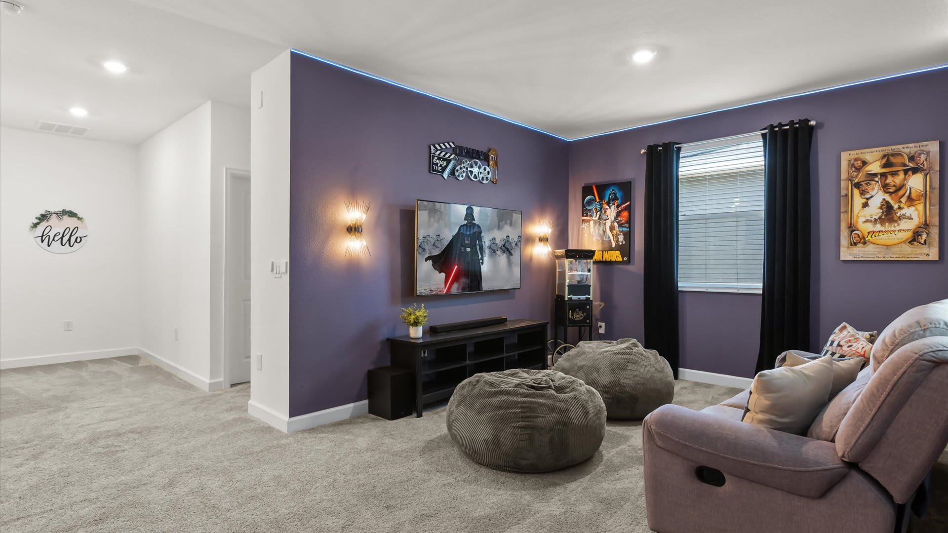A cozy media room featuring a large wall-mounted TV displaying a Star Wars scene, flanked by two modern sconces, against a purple wall with blue LED strip lighting. A black media console sits below the TV, accompanied by two grey bean bag chairs and a grey reclining sofa.