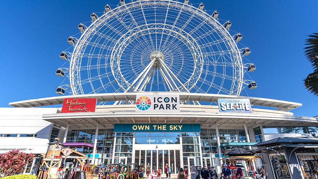 The iconic ICON Park complex under a clear blue sky, featuring a towering white Ferris wheel, the main entrance with 'OWN THE SKY' signage, and various attractions like Madame Tussauds and SEA LIFE Orlando.