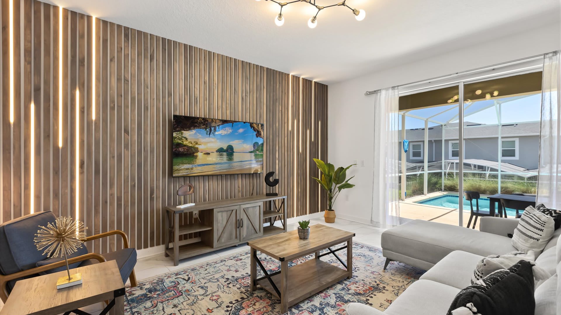 A contemporary living room features a large wall with vertical wooden slats and integrated LED lighting, showcasing a wall-mounted TV. A light gray sectional sofa and a wooden coffee table with a patterned rug complete the space, with sliding doors opening to a pool area.