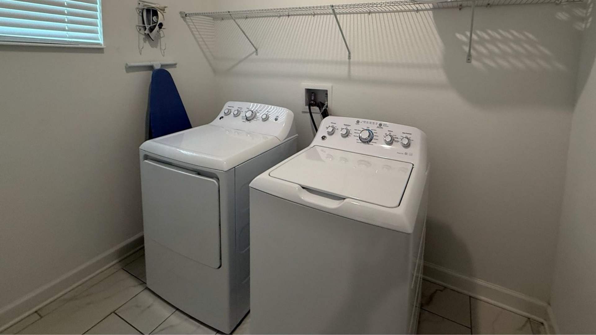 Functional laundry room featuring a white top-loading washing machine and a white front-loading dryer. A wire shelf is mounted above, and an ironing board hangs on the wall.