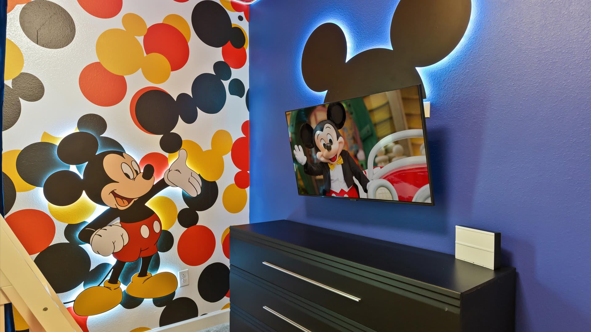Close-up of a vibrant Mickey Mouse themed wall with illuminated character cutouts, a wall-mounted television, and a dark dresser. The blue wall contrasts with the colorful polka dot pattern.