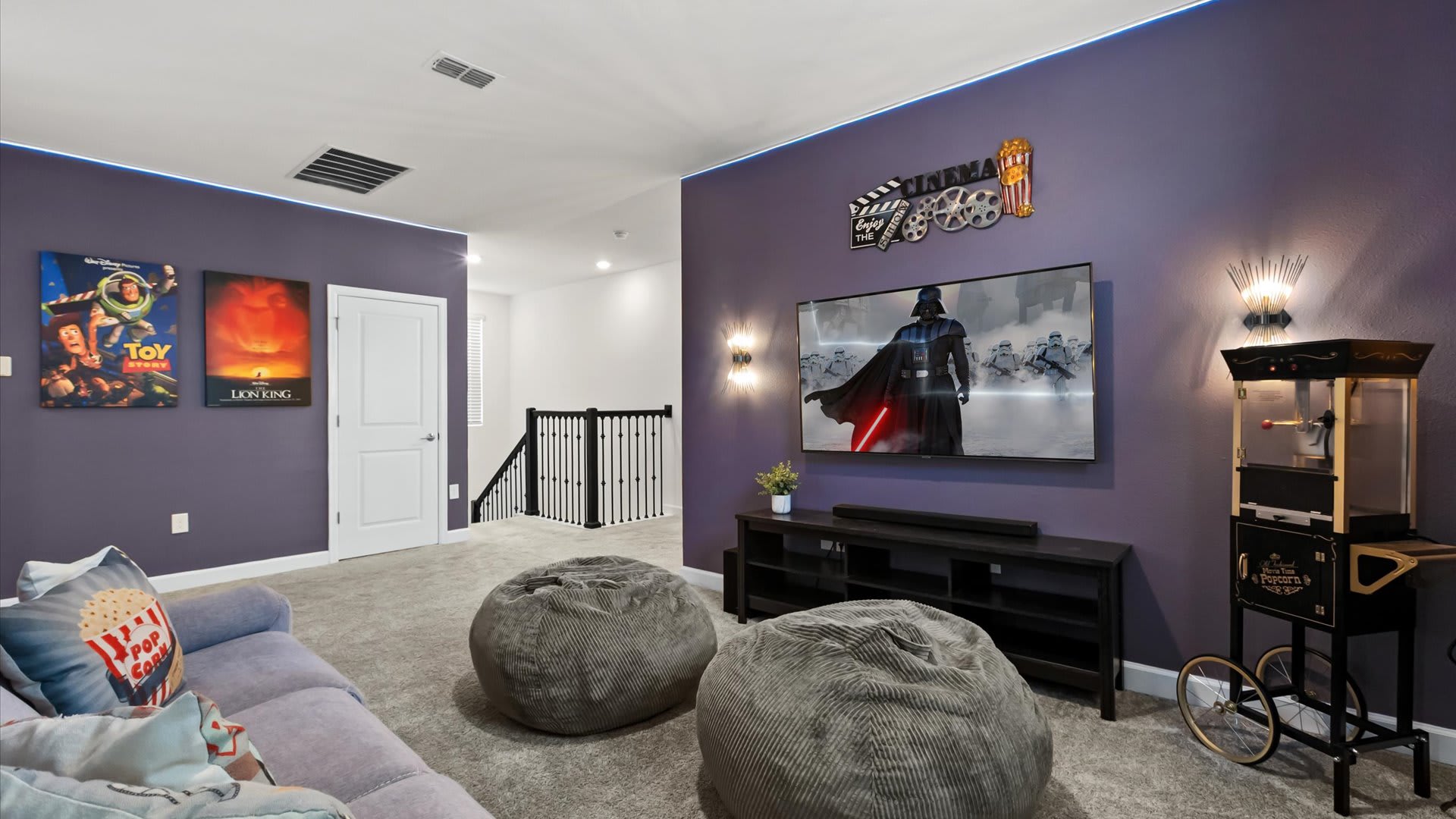 A spacious media room with purple walls and blue LED strip lighting, featuring a large wall-mounted TV, a black media console, and two grey bean bag chairs on a light grey carpet. Movie posters for Toy Story and The Lion King adorn a white wall, and a vintage popcorn machine stands in the corner.