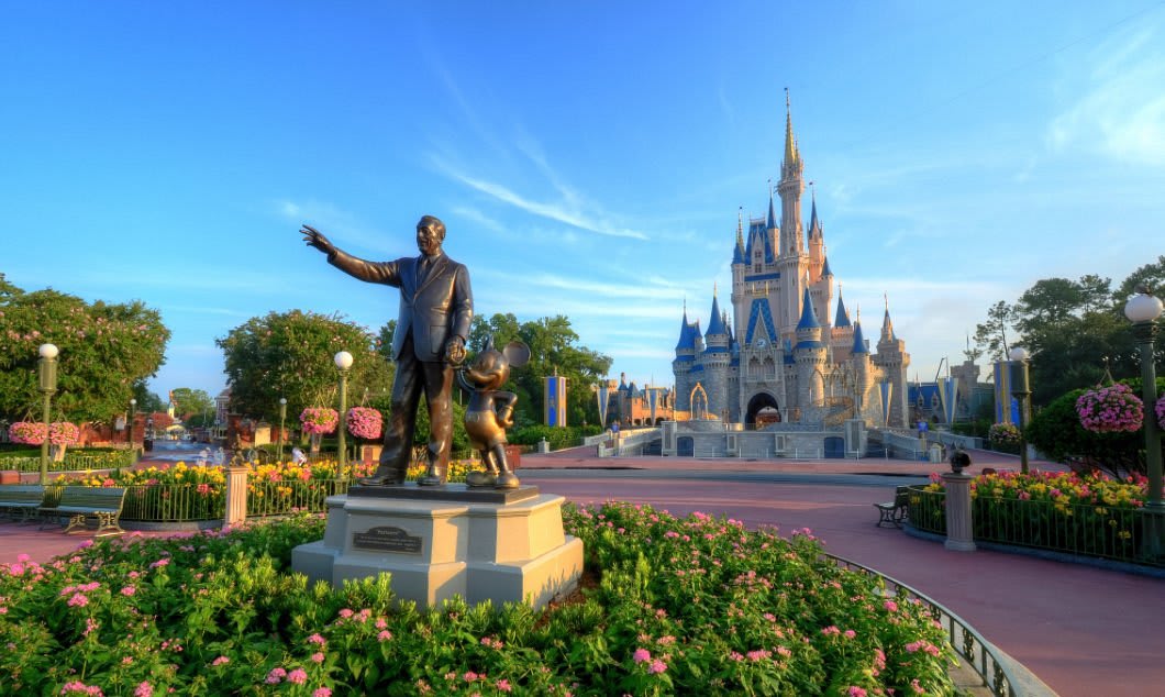 Iconic Cinderella's Castle at Walt Disney World, with the 'Partners' statue of Walt Disney and Mickey Mouse in the foreground. The area is adorned with vibrant pink flowers and lush greenery under a clear blue sky.