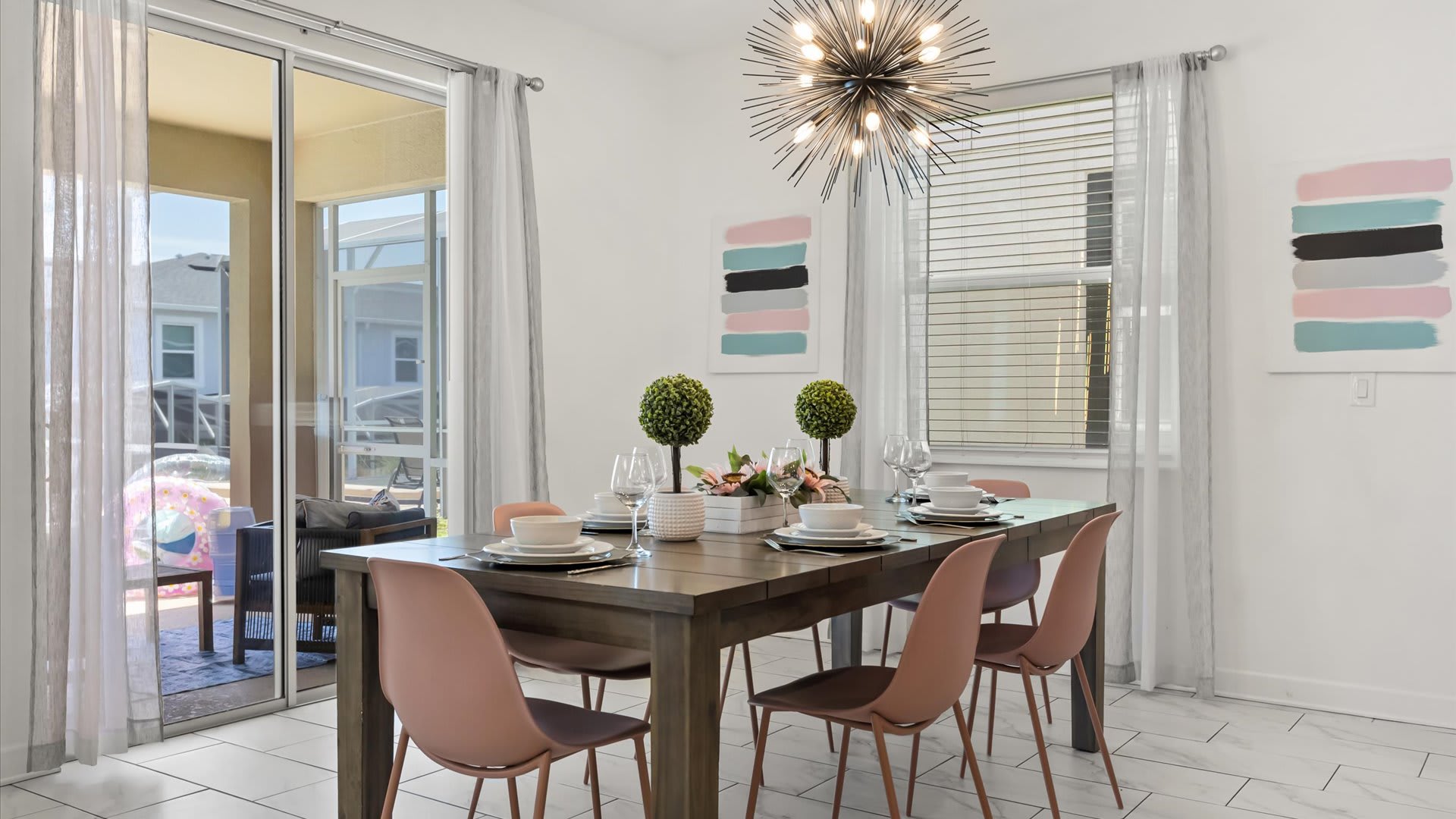 A modern dining room features a dark wood rectangular table set with white dishes and two small topiary plants, surrounded by six blush pink chairs. Large sliding glass doors lead to an outdoor patio, and a sputnik-style chandelier hangs above.