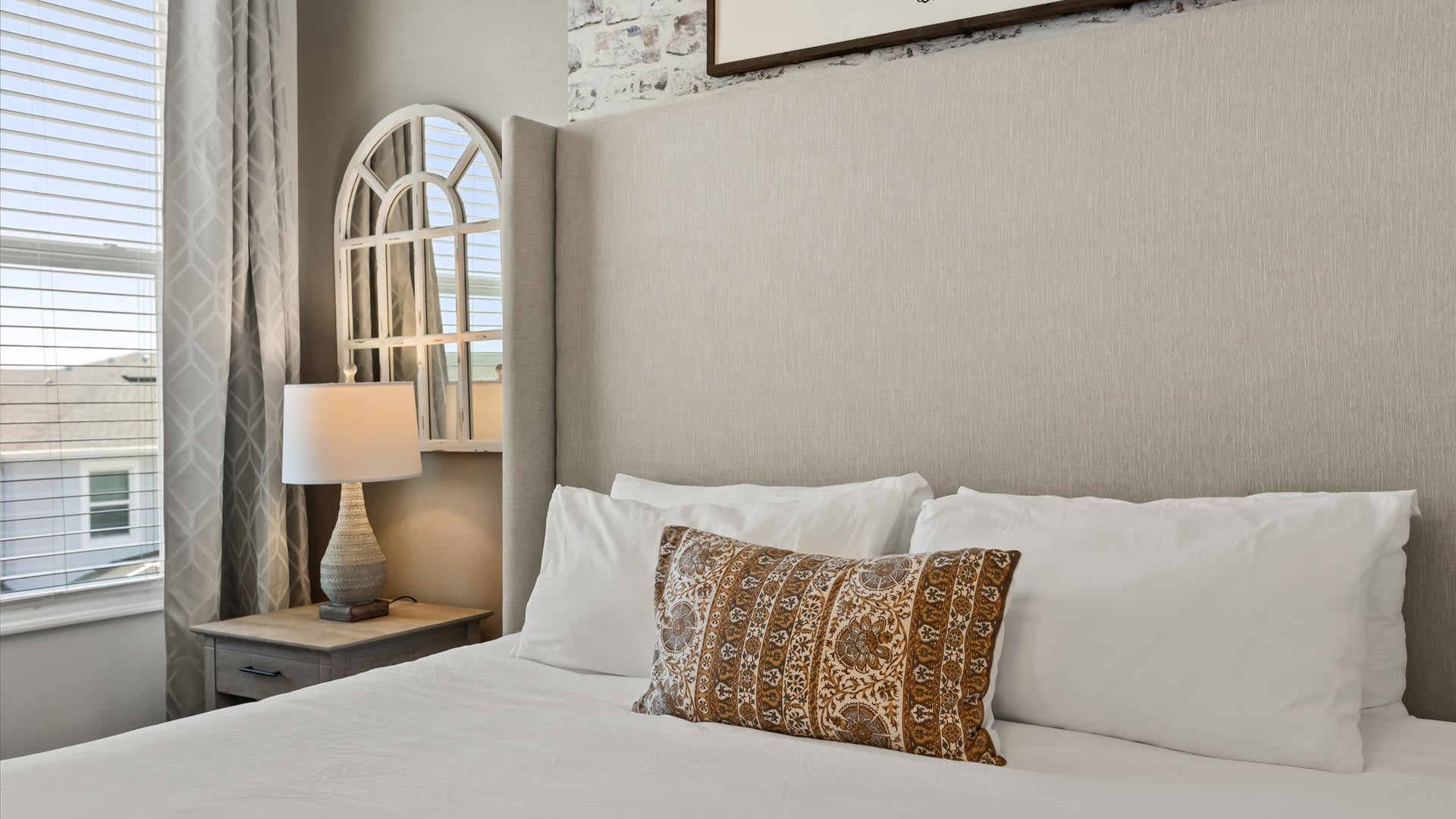 A close-up of the comfortable king-sized bed in bedroom-1, featuring a tall, light beige upholstered headboard and crisp white bedding with a patterned accent pillow. A rustic nightstand with a lamp and an arched mirror are visible beside the bed.