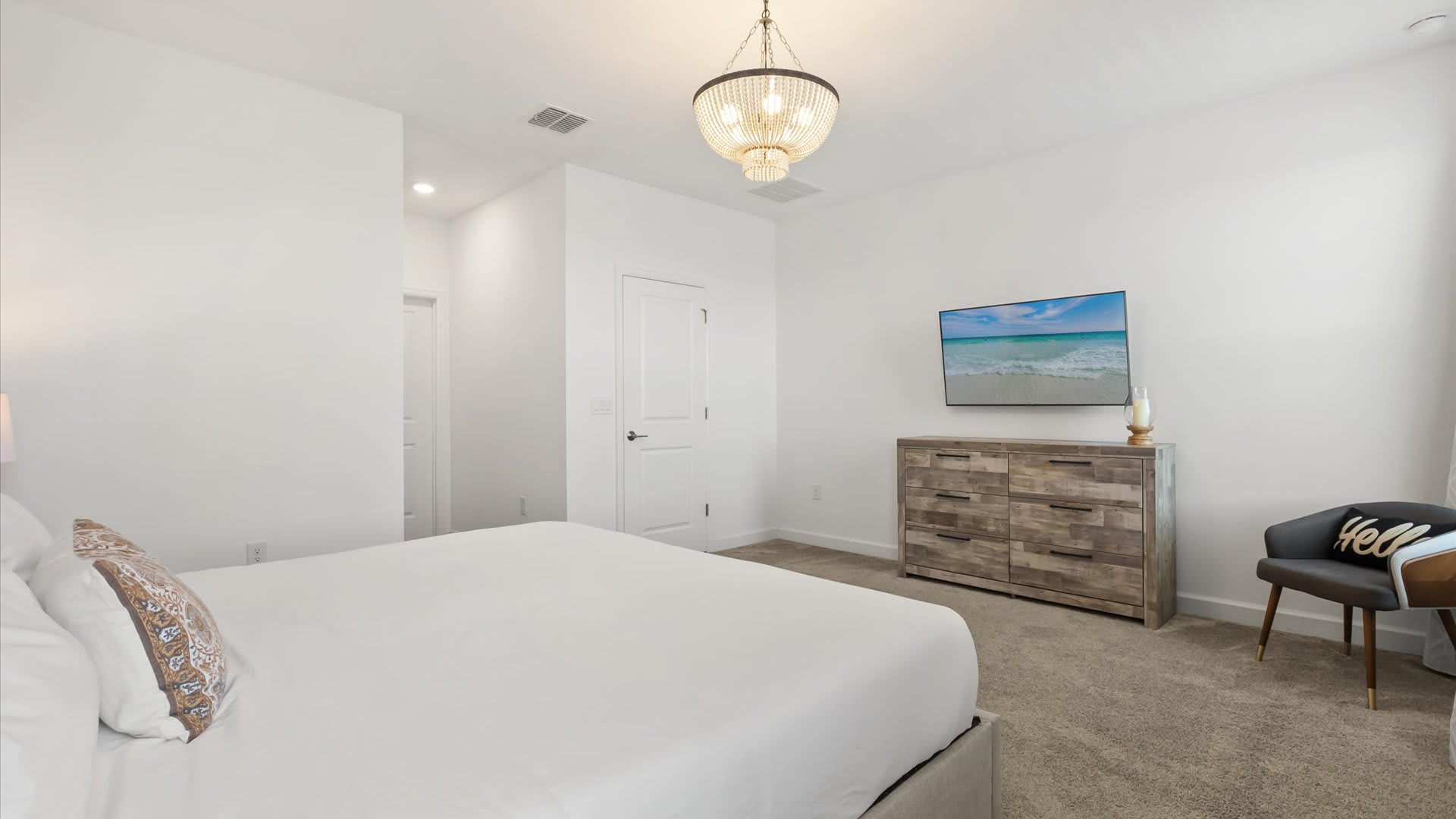 A spacious bedroom with white walls and light grey carpet, featuring a large bed with a tall upholstered headboard and a rustic wooden dresser with a wall-mounted TV. A beaded chandelier illuminates the room, and a dark accent chair with a "hello" pillow adds a welcoming touch.