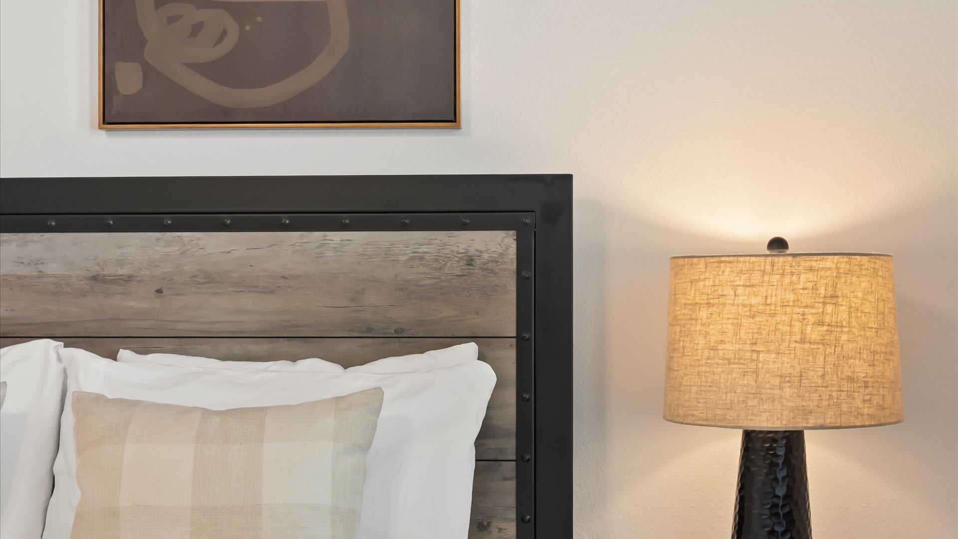 Close-up view of the rustic wood and metal headboard with white pillows and a plaid accent pillow, alongside a nightstand with a textured table lamp. A framed artwork hangs above the bed.