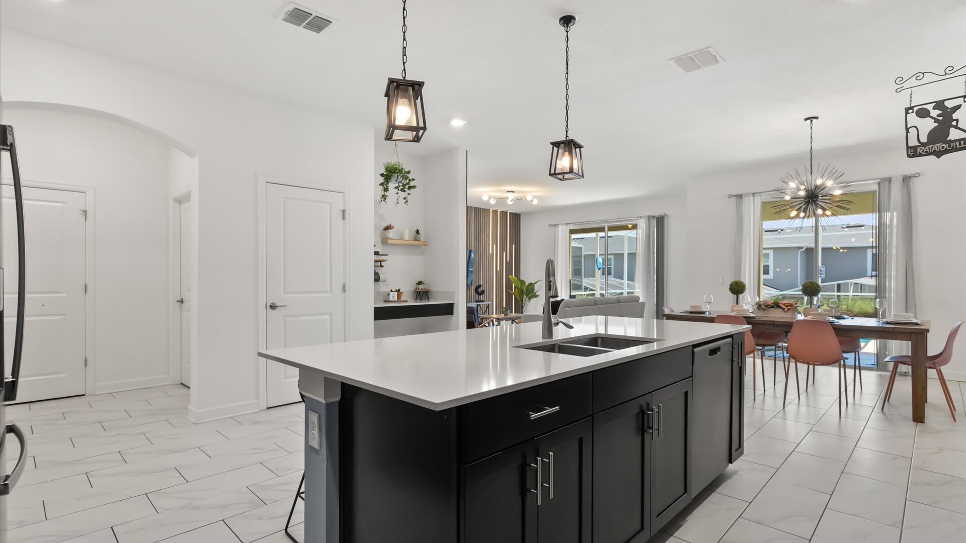 This kitchen view emphasizes the large central island with a double sink and black base, contrasting with the white tile floor. The open layout connects to the dining area and living room, with black pendant lights hanging above the island and a "La Ratatouille" sign visible.