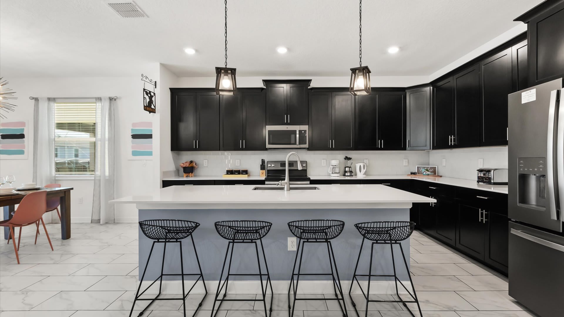 A spacious, open-concept kitchen showcases extensive black cabinetry, white countertops, and a large island with four black wire bar stools. Stainless steel appliances are integrated, and the dining area with its modern chandelier is visible in the background, connecting the spaces.