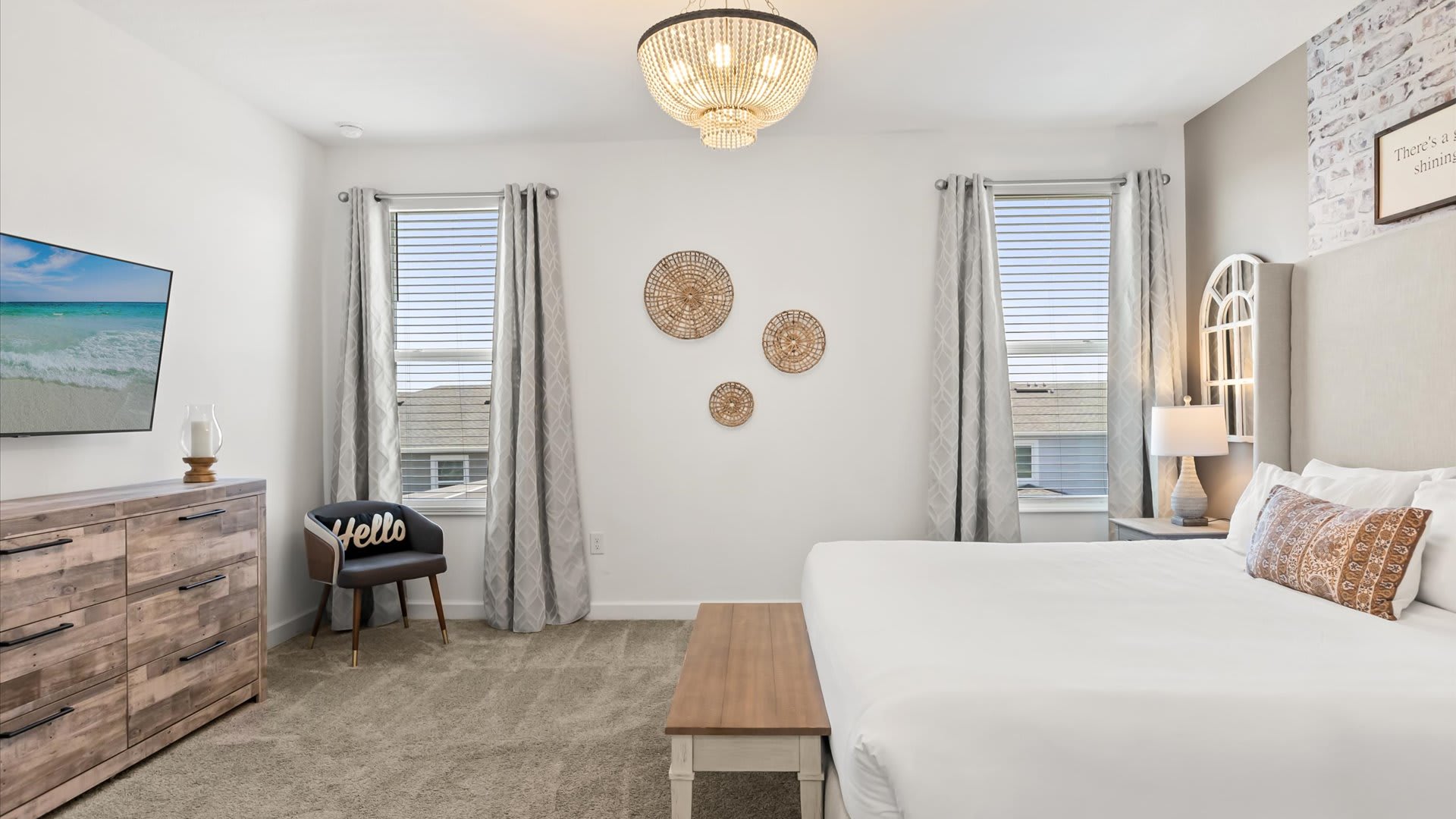 A bright and airy bedroom featuring a large bed with a tall, upholstered headboard, a wooden bench at the foot, and a rustic wooden dresser with a TV mounted above it. Two windows with light grey curtains and blinds allow natural light, while a beaded chandelier hangs from the ceiling.