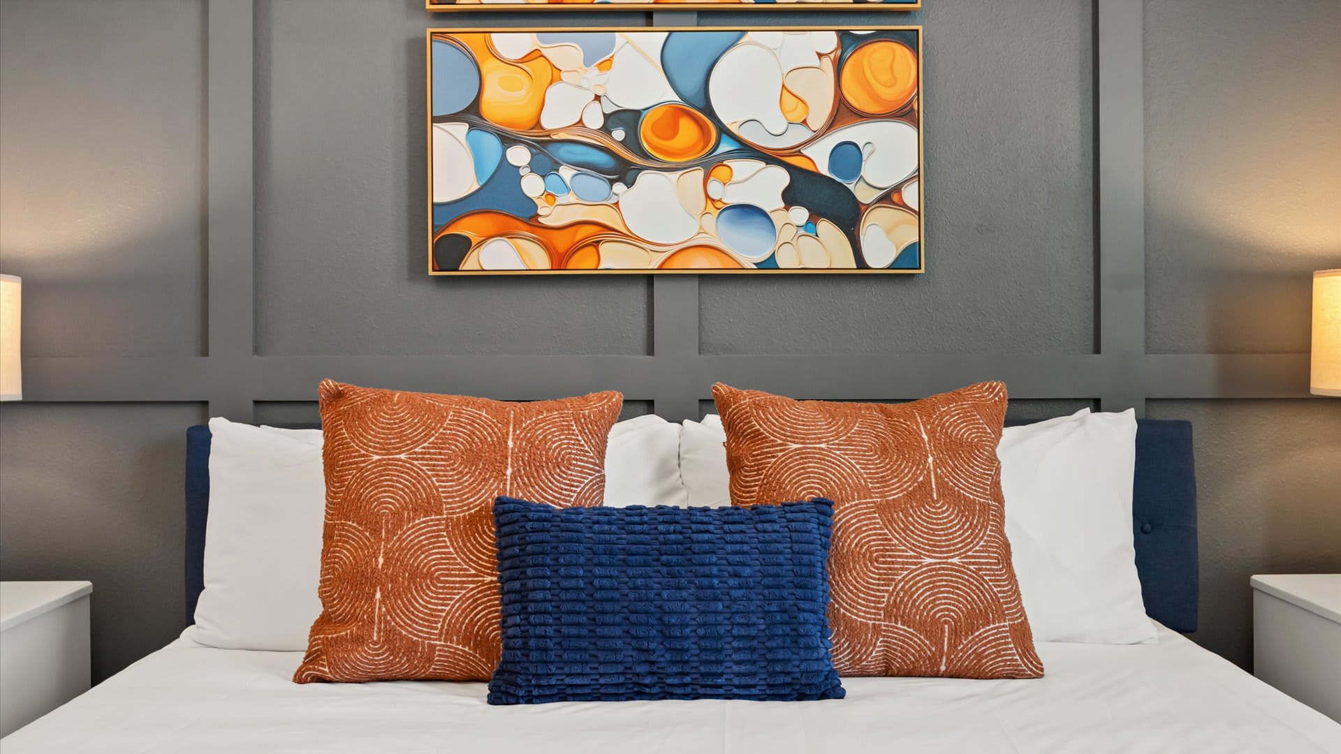 A close-up of the bed in bedroom-2, showcasing a blue upholstered headboard against a dark grey paneled wall, adorned with white bedding, two orange patterned pillows, and a blue textured lumbar pillow. Two abstract art pieces hang above the bed.