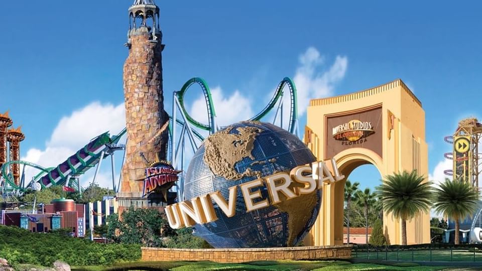 An aerial view of Universal Studios Florida, featuring the iconic Universal globe, the Islands of Adventure lighthouse, and various roller coasters under a bright blue sky.