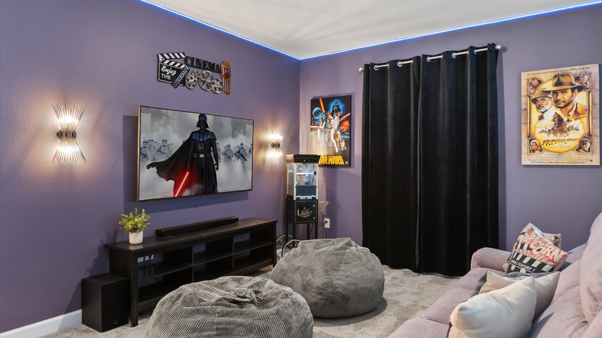 A vibrant media room with purple walls, blue LED strip lighting, and a large wall-mounted TV displaying a Star Wars scene. A vintage popcorn machine and a Star Wars poster are visible, alongside a grey reclining sofa and two grey bean bag chairs on a light grey carpet.