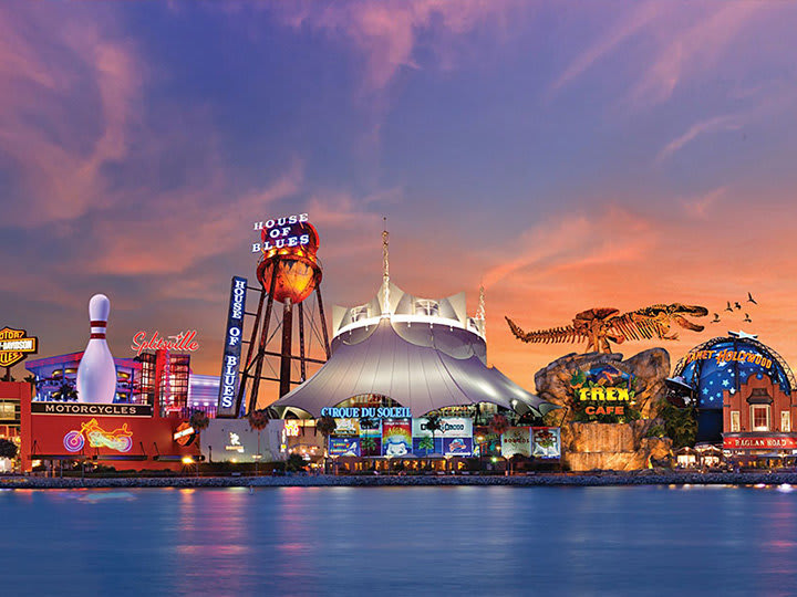 A panoramic view of a vibrant entertainment complex at dusk, featuring various themed restaurants and venues like House of Blues, Cirque du Soleil, and T-Rex Cafe, illuminated against a colorful sunset sky.
