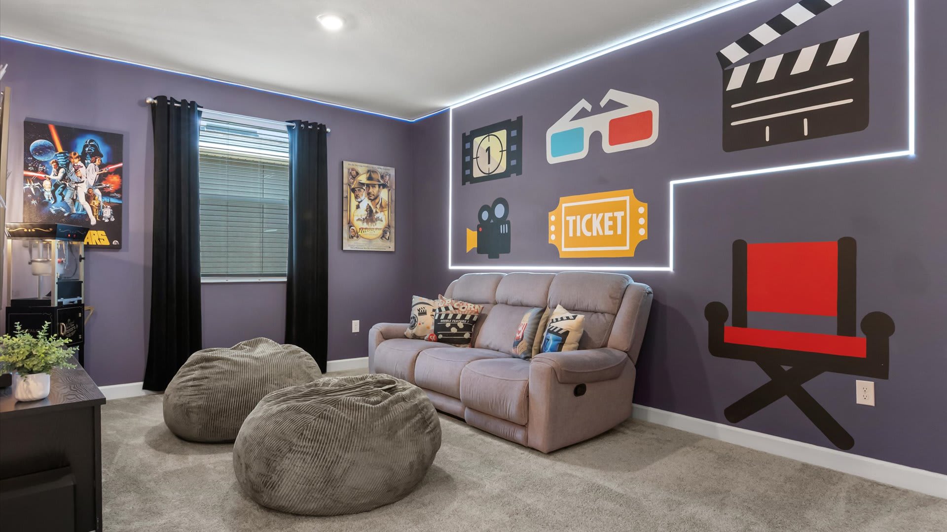 A vibrant media room with purple walls adorned with movie-themed decals like a clapperboard, 3D glasses, and a director's chair, illuminated by white LED strip lighting along the ceiling and walls. Two grey bean bag chairs and a grey reclining sofa provide seating on a light grey carpet.