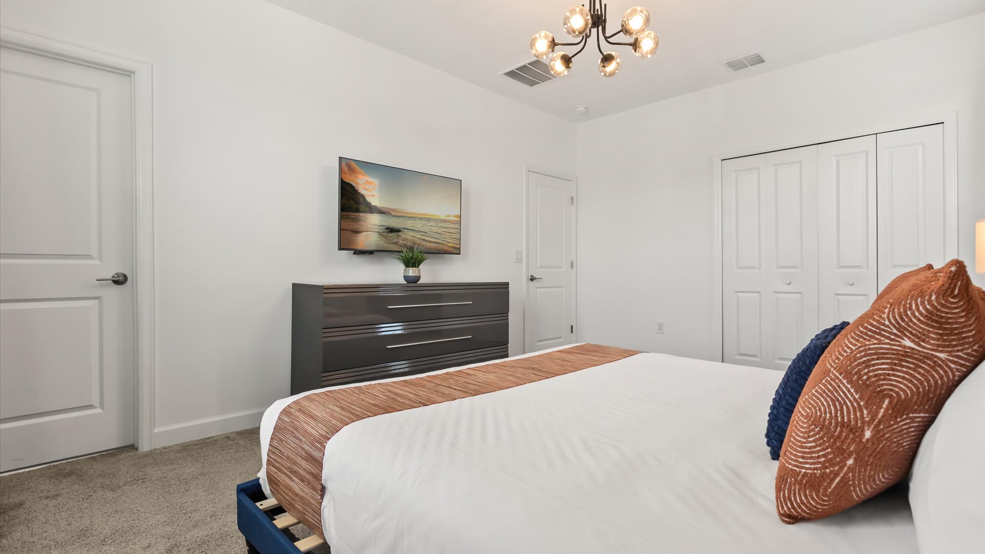 Bright bedroom with white walls, a dark gray dresser topped with a TV, and a bed featuring white linens and decorative orange and blue pillows. A modern sputnik-style chandelier hangs from the ceiling.
