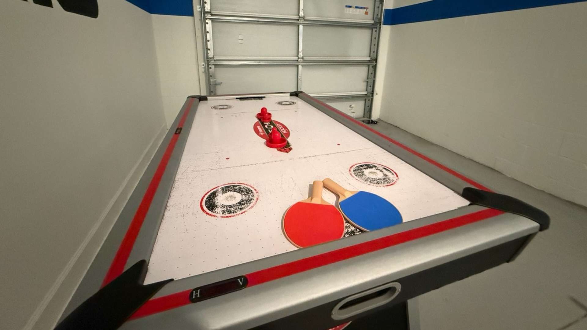 A bright game room features an air hockey table with red and blue paddles, set against a white wall with a blue stripe and a garage door in the background. The table has a silver and red trim, and the floor is light-colored.
