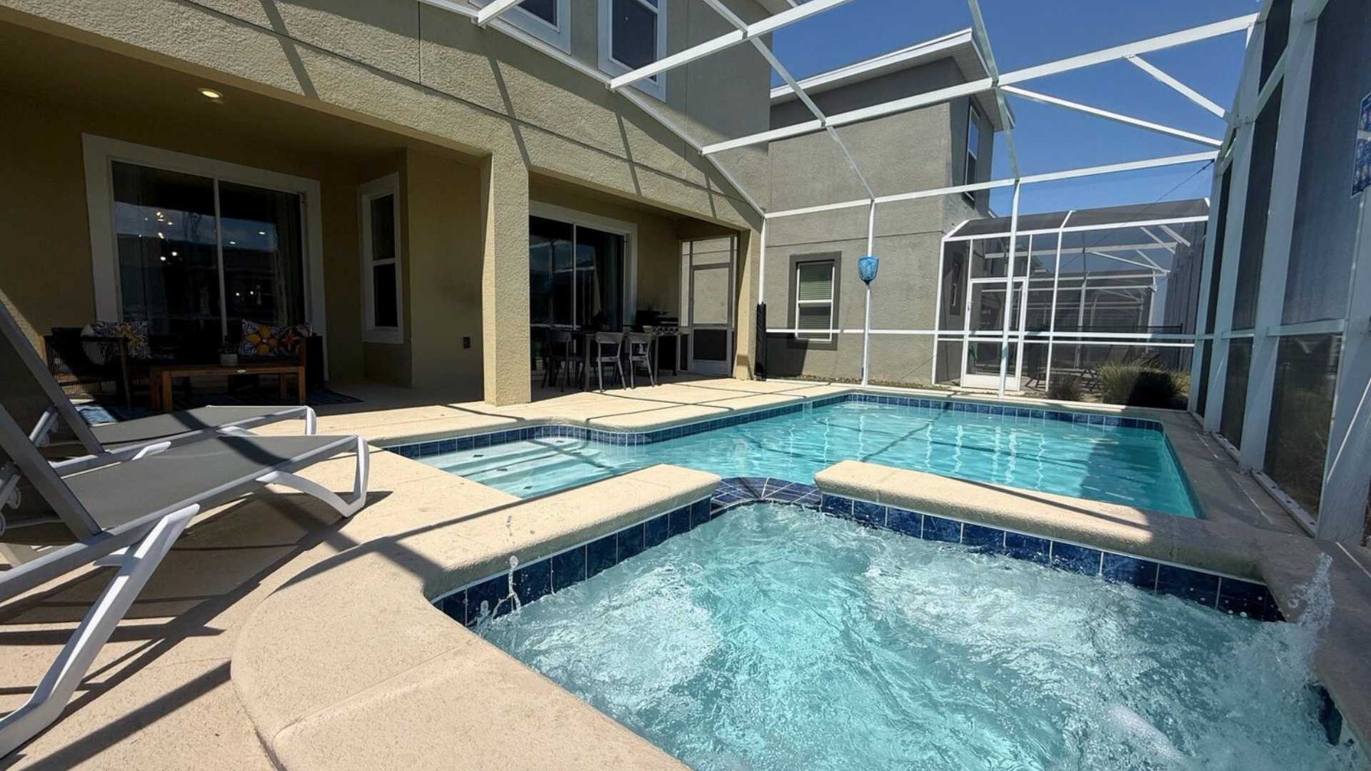 Screened-in outdoor pool area with a rectangular swimming pool and an attached hot tub, surrounded by a concrete deck and lounge chairs, with a covered patio area visible.