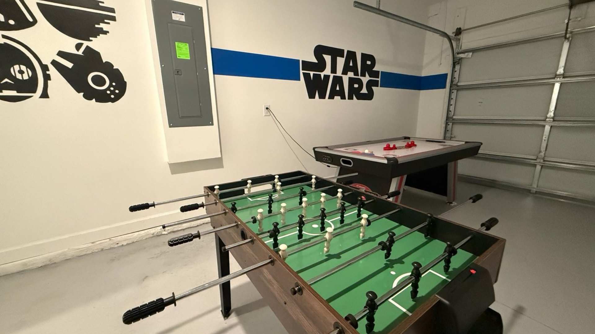 Garage converted into a game room with a foosball table and an air hockey table, featuring Star Wars themed wall decor and a blue and white stripe accent.